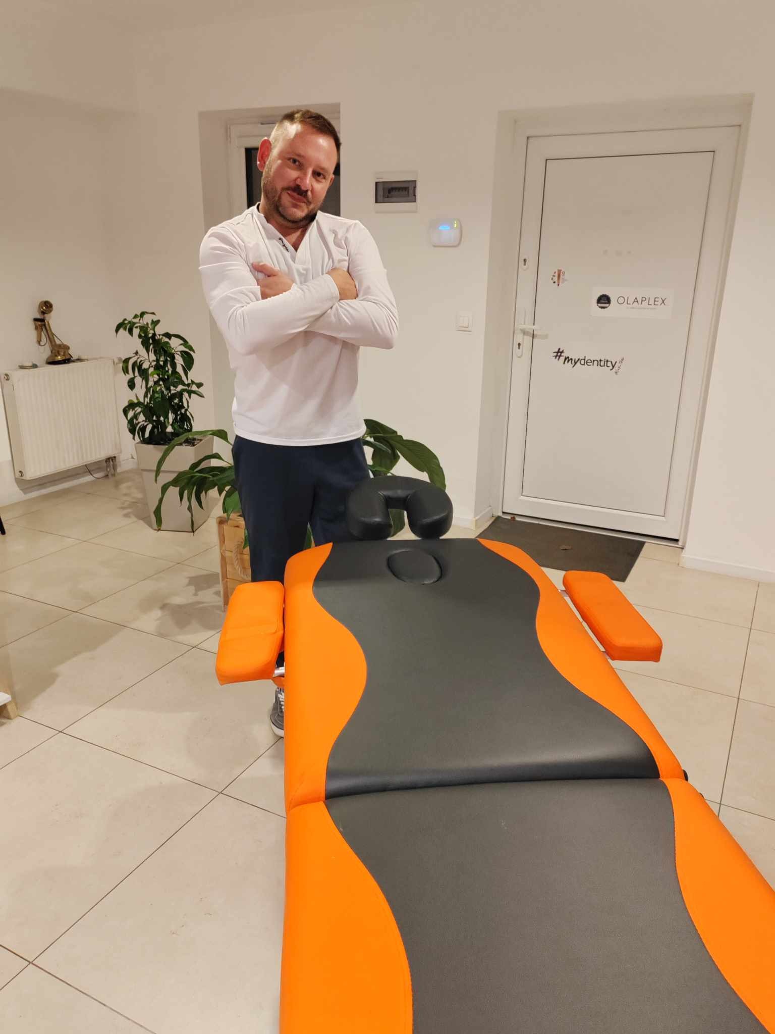 Mężczyzna w białej koszuli stoi obok pomarańczowo-szarego stołu do masażu w jasnym pomieszczeniu z roślinami i drzwiami z napisem Olaplex #myidentity.