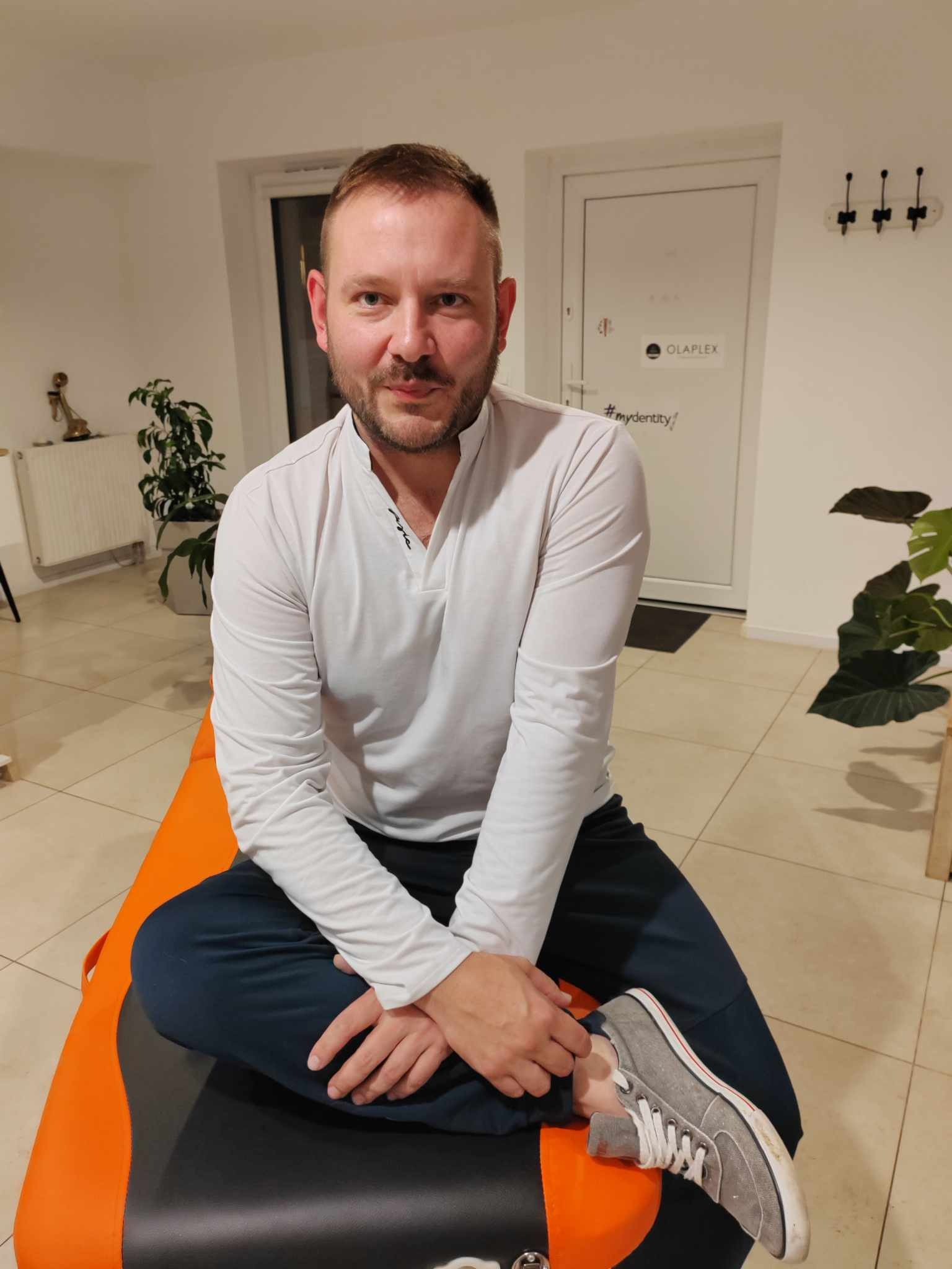 Mężczyzna w białej bluzce siedzi ze skrzyżowanymi nogami na pomarańczowym stole do masażu w jasnym pomieszczeniu z roślinami doniczkowymi.