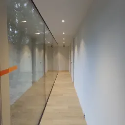 Długi korytarz z jasną drewnianą podłogą, białymi ścianami i dużym oknem z widokiem na drzewo. Czarne, nowoczesne lampy sufitowe. Minimalistyczny design.