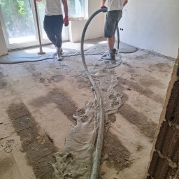 Tech-mal.pl - Wylewka samopoziomująca rozprowadzana w pomieszczeniu z oknami. Widoczny wąż doprowadzający masę i pracownicy w gumowych butach. Laserowy niwelator wskazuje poziom.