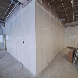 Tech-mal.pl - Wnętrze w trakcie prac: ściany po nałożeniu gładzi, widoczne instalacje pod sufitem, surowy beton na podłodze, przygotowane do dalszych prac wykończeniowych.