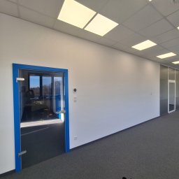 Tech-mal.pl - Białe ściany biura z szarą wykładziną i niebieskimi ramami szklanych drzwi. Widoczne oświetlenie sufitowe i gniazdka elektryczne. Minimalistyczny design.