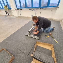 Tech-mal.pl - Montaż paneli podłogowych w biurze: mężczyzna docina panel specjalistycznym narzędziem, obok leżą panele w kartonie i miarka. W tle widać innego pracownika.