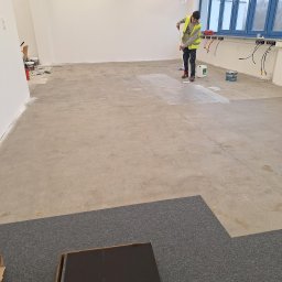 Tech-mal.pl - Pracownik w kamizelce maluje podłogę wałkiem w jasnym pomieszczeniu z dużymi oknami. Widoczne puszki z farbą i narzędzia malarskie. Częściowo położona wykładzina podłogowa.