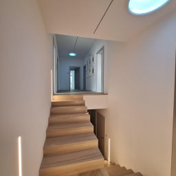 Tech-mal.pl - Nowoczesne schody drewniane z subtelnym oświetleniem LED wzdłuż ścian, prowadzące do jasnego korytarza z okrągłym świetlikiem sufitowym i minimalistycznymi dekoracjami.