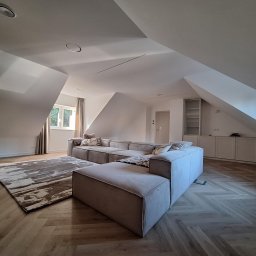 Tech-mal.pl - Jasny salon na poddaszu z beżową sofą modułową, wzorzystym dywanem i parkietem w jodełkę. Okno z zasłonami w tle. Minimalistyczny design.