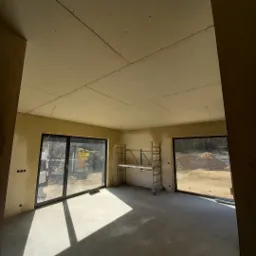 Wnętrze w trakcie montażu płyt karton-gips na suficie, widoczne szpachlowane ściany, aluminiowy ruszt i duże okna z widokiem na zewnątrz.