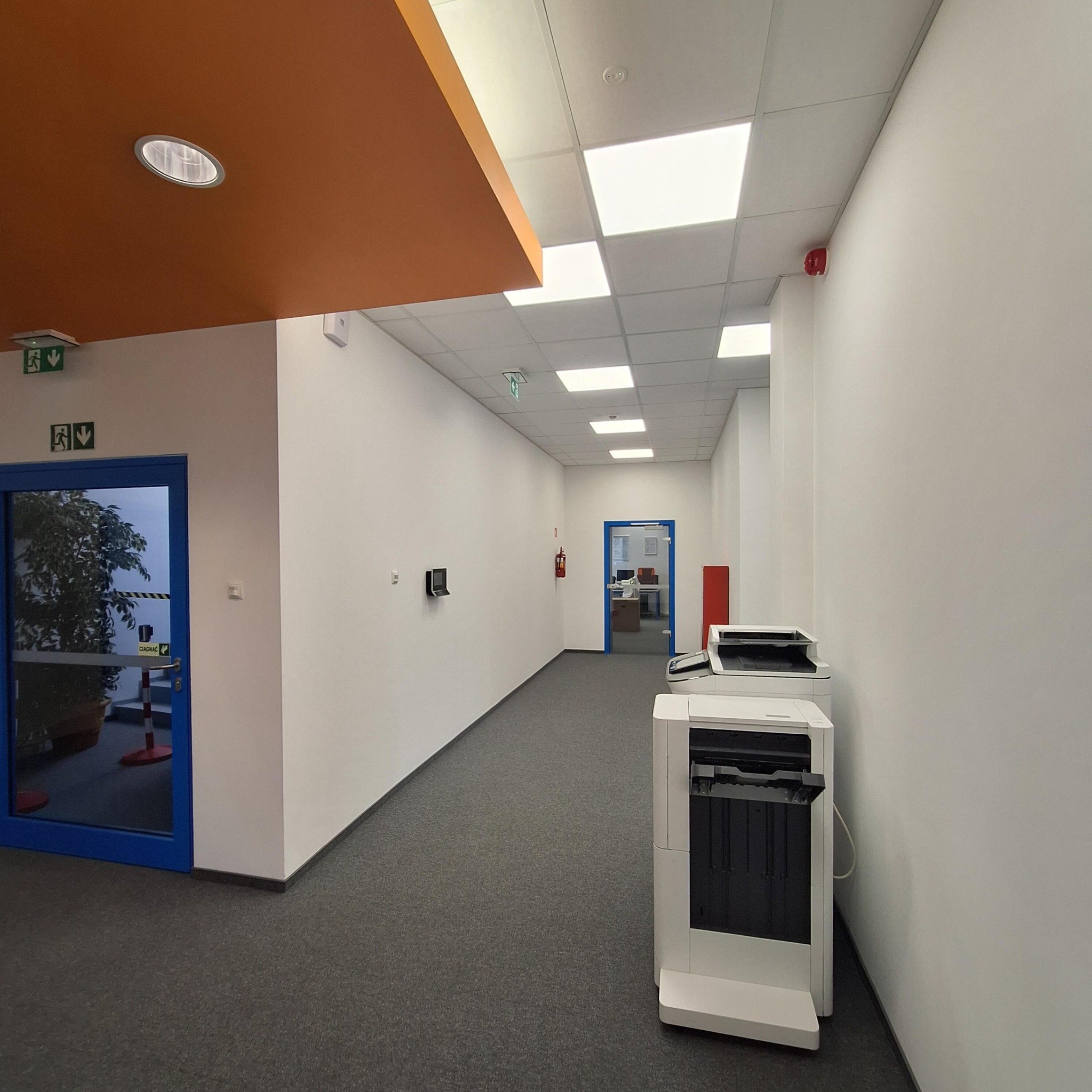 Korytarz biurowy z szarą wykładziną, białymi ścianami i sufitem kasetonowym. Widoczne drzwi z rośliną, drukarka i pomarańczowy element sufitu.