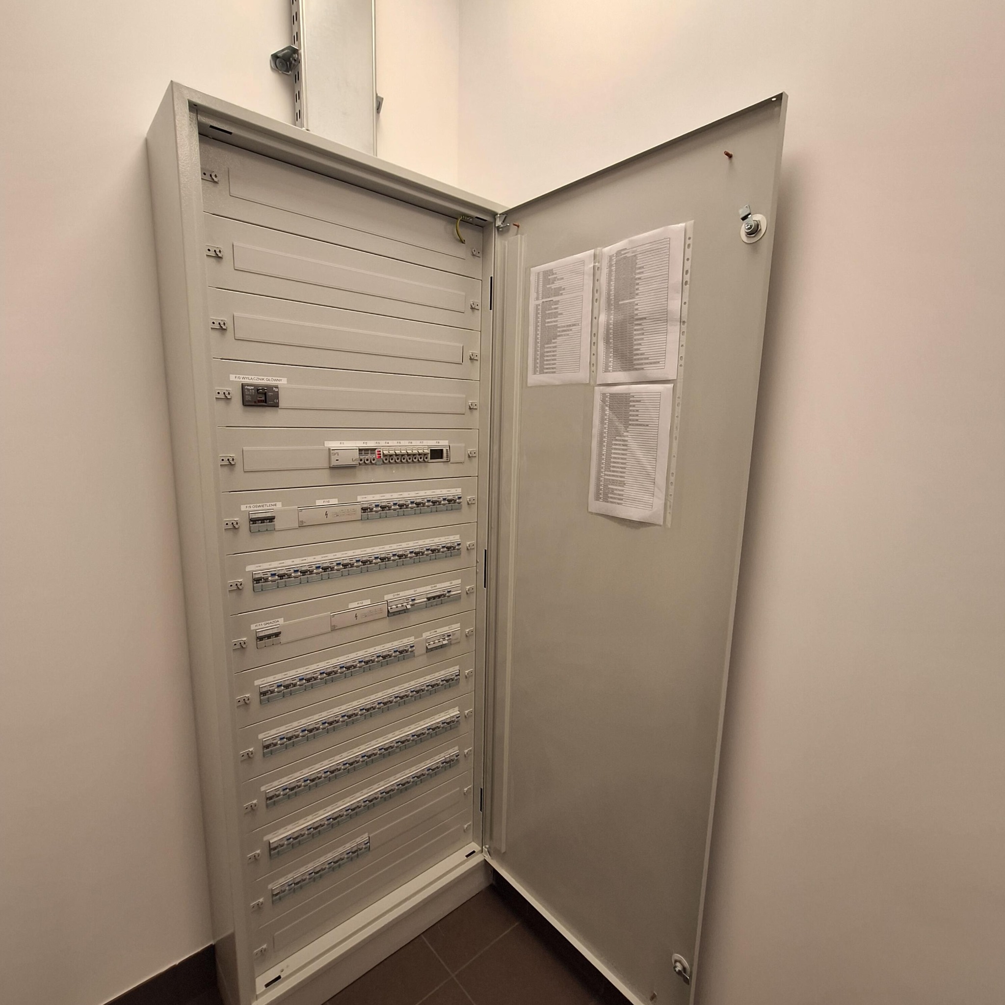 Otwarta, szara szafa elektryczna z listwami i wyłącznikami, zamontowana na ścianie w jasnym pomieszczeniu. Na drzwiach szafy widoczne dokumenty z listą.