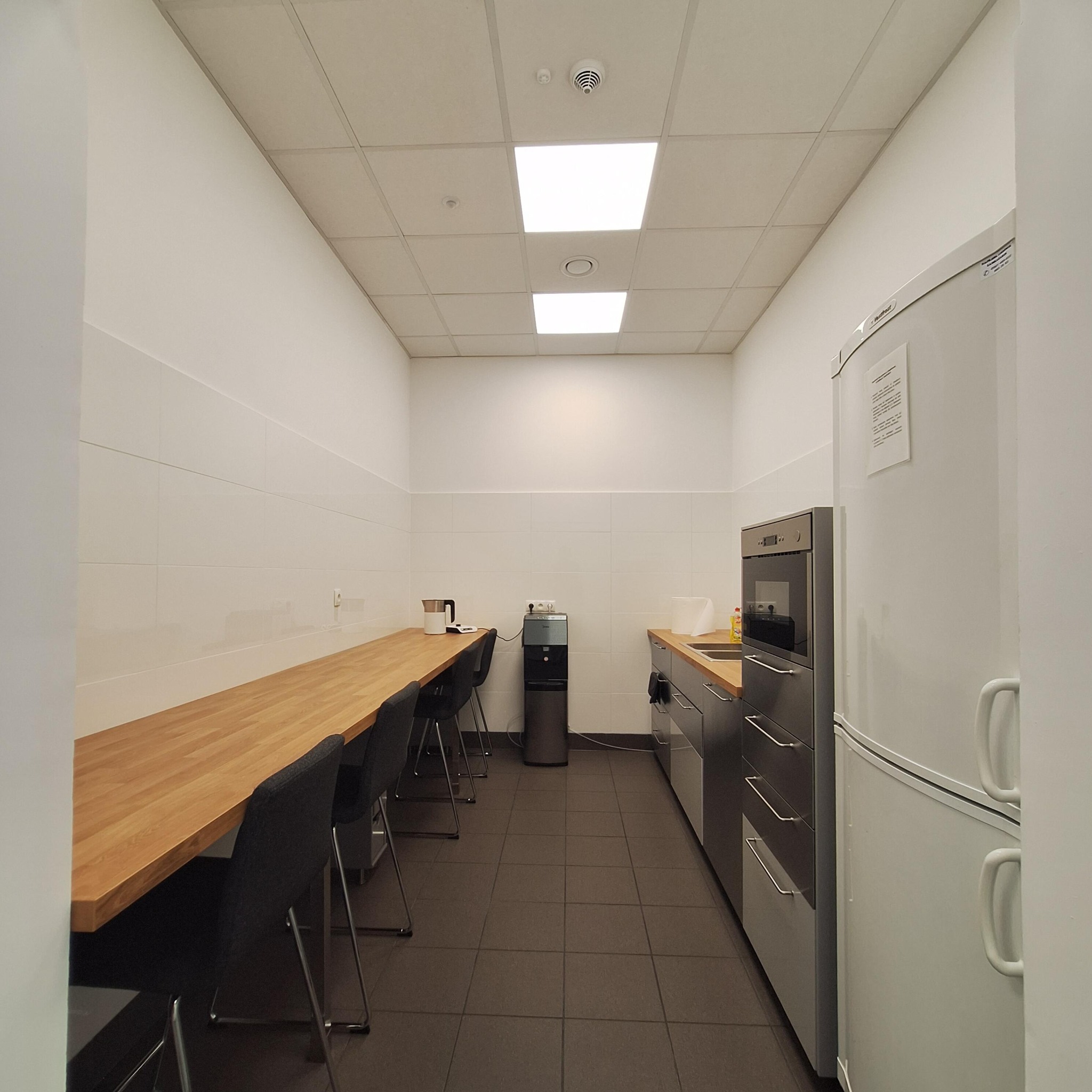 Jasne biuro z długim drewnianym blatem, krzesłami, aneksem kuchennym i białymi ścianami wykończonymi płytkami. Widok na przestrzeń socjalną w biurze.