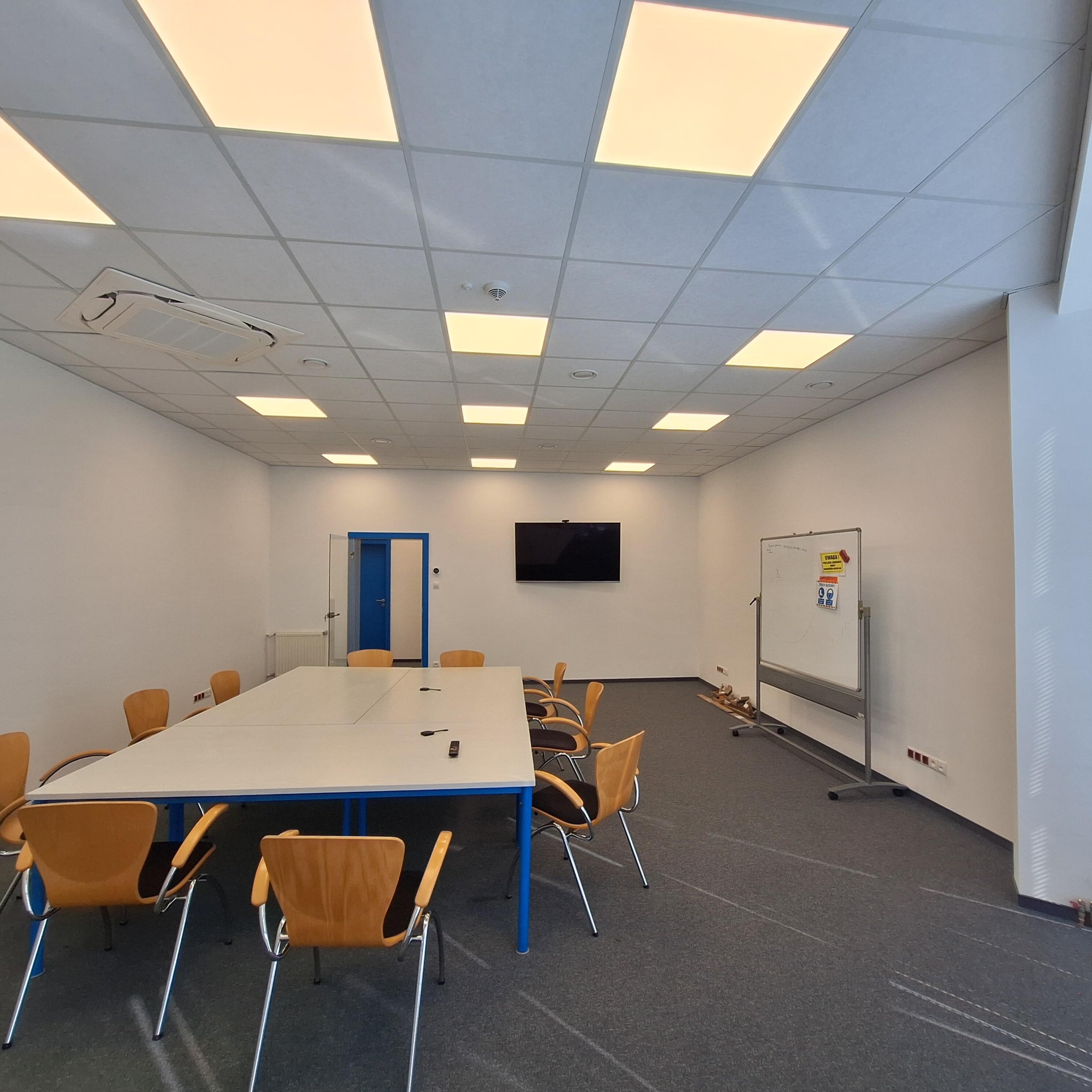 Jasne biuro po remoncie: białe ściany, sufit kasetonowy z oświetleniem LED, szary dywan, stół konferencyjny z krzesłami. Widoczny telewizor i tablica suchościeralna.