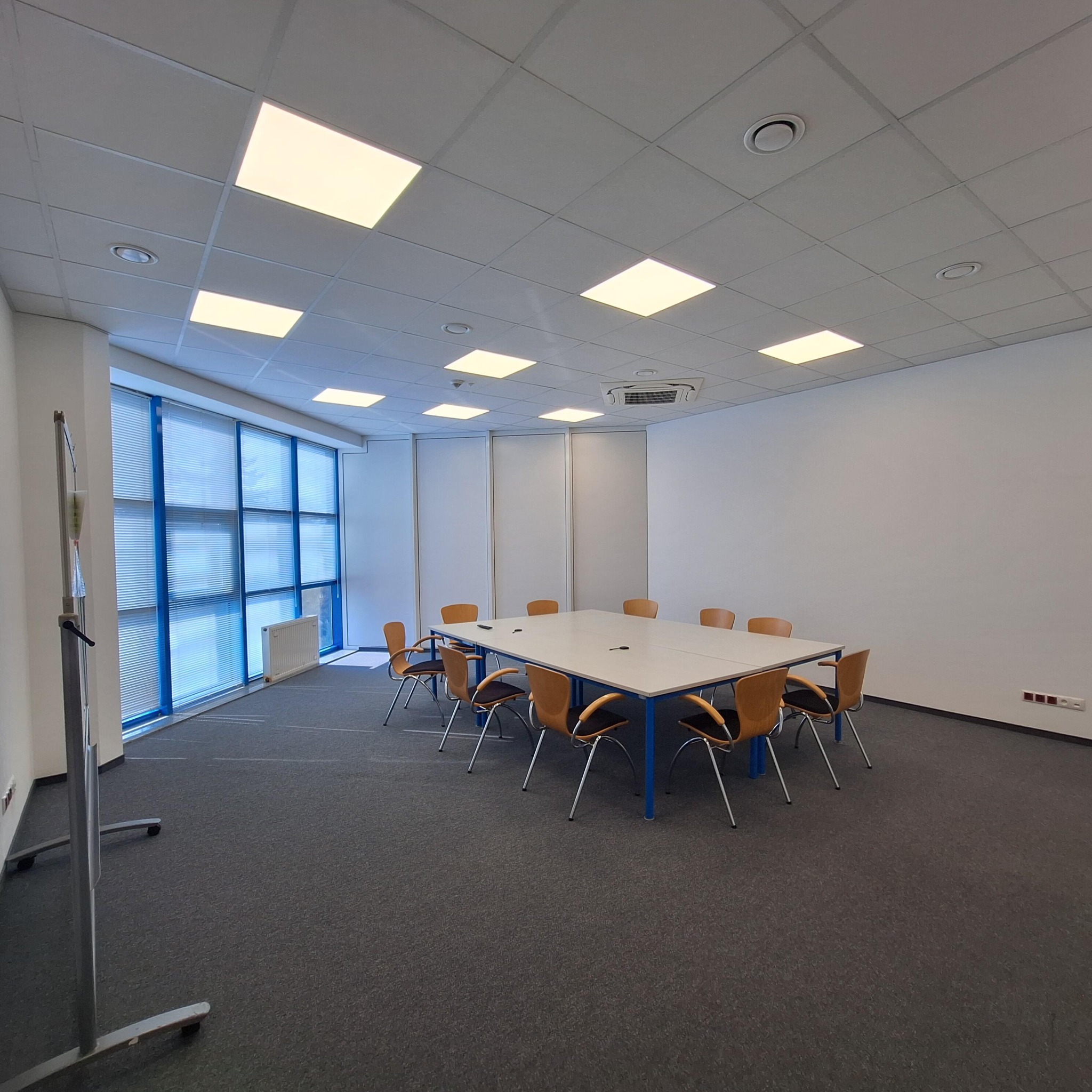 Jasne biuro po remoncie: białe ściany, sufit kasetonowy, szara wykładzina, stół konferencyjny z krzesłami. Duże okno z żaluzjami. Minimalistyczny, nowoczesny design.