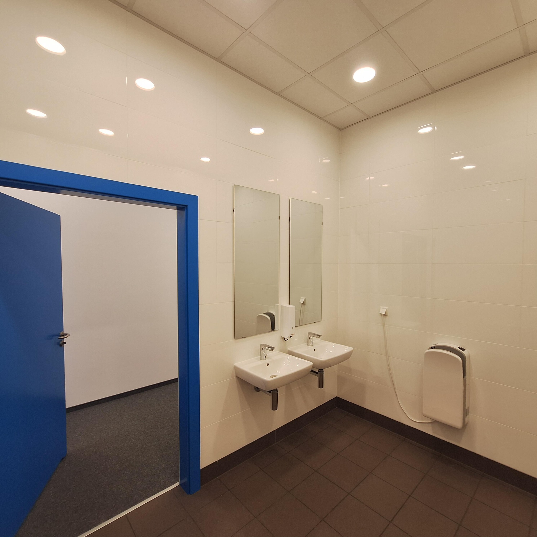 Wykończona łazienka w biurze z dwoma umywalkami, lustrami i suszarką do rąk. Białe płytki na ścianach, brązowe na podłodze. Niebieskie drzwi otwarte do innego pomieszczenia.