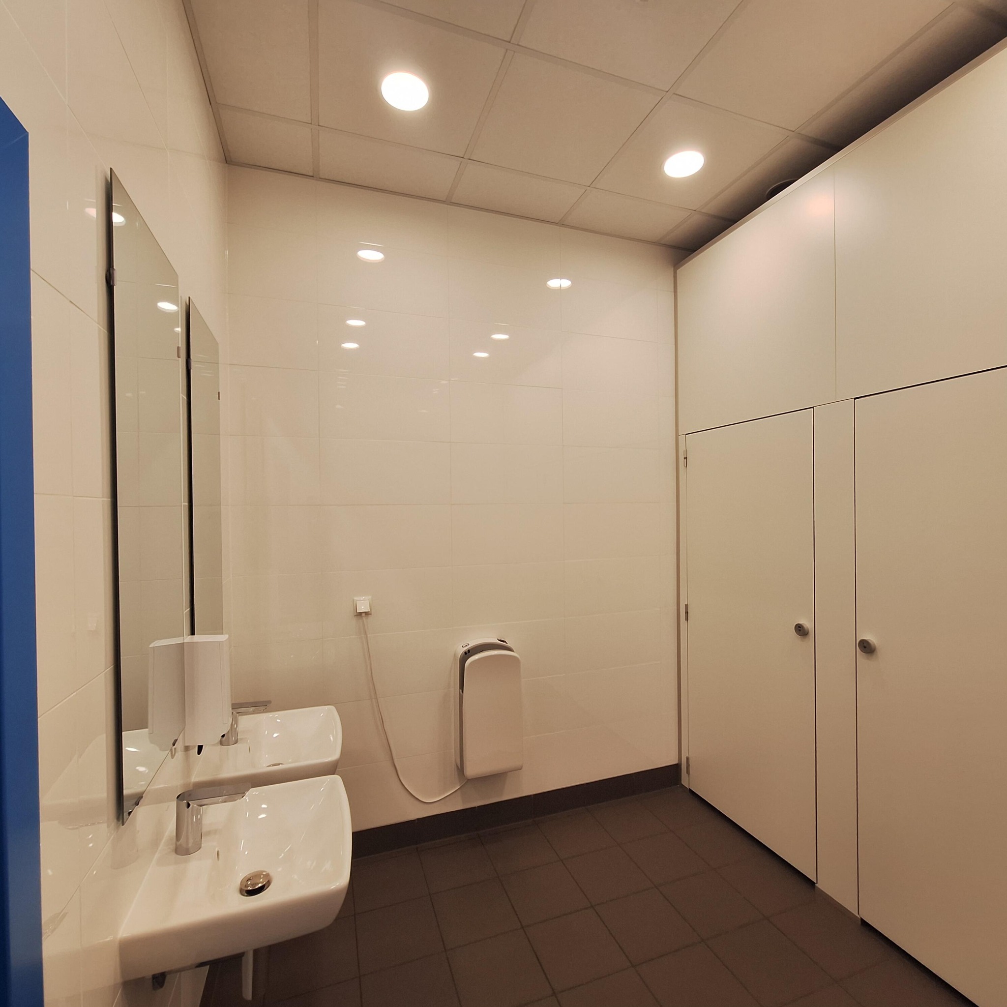 Wykończona łazienka w biurze: białe płytki, dwie umywalki z lustrami, suszarka do rąk i szafki w zabudowie. Minimalistyczny design, oświetlenie punktowe.