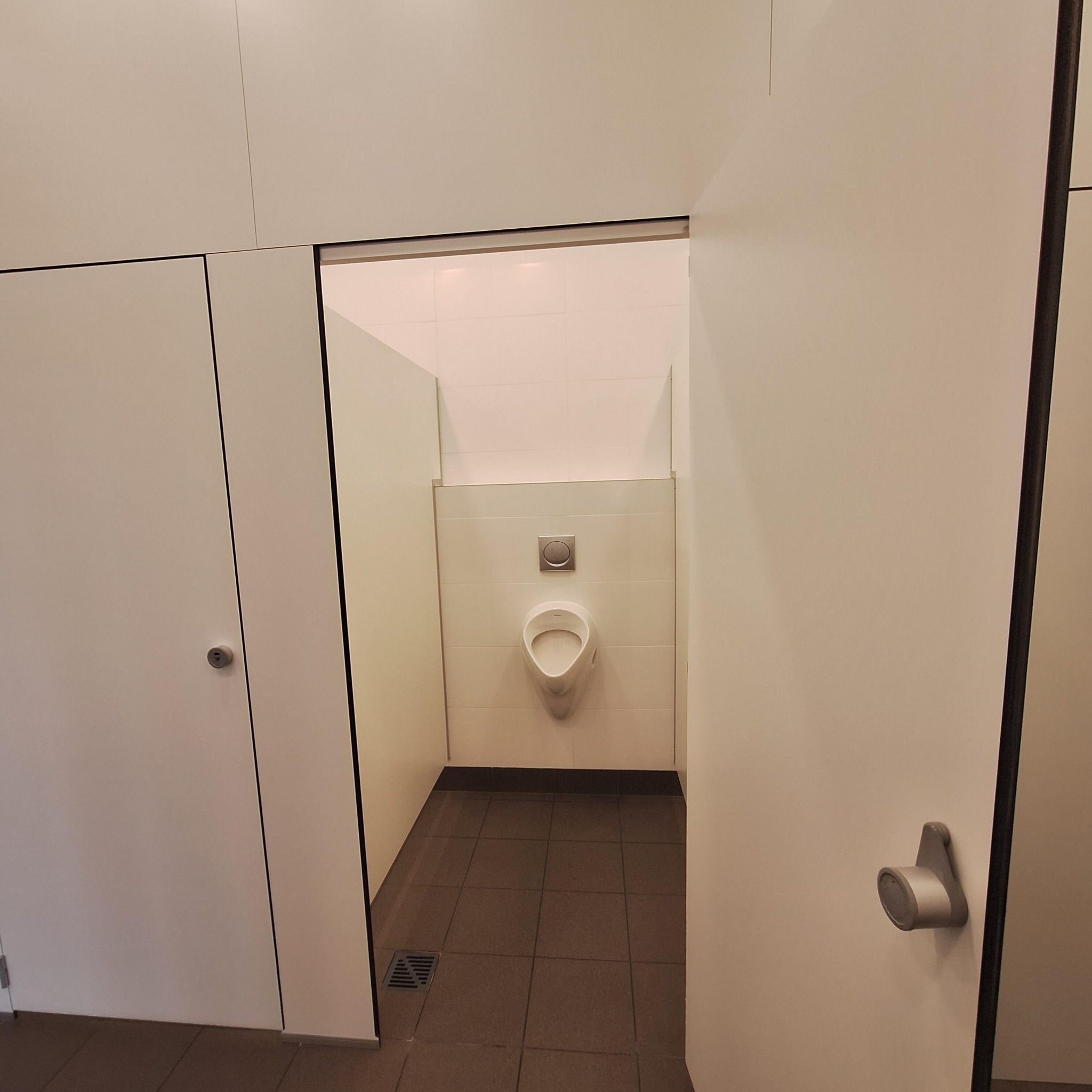 Minimalistyczna toaleta publiczna z białymi ścianami i brązowymi płytkami podłogowymi, widziana z perspektywy wejścia. Uwagę przykuwa nowoczesny pisuar na tle białych płytek.