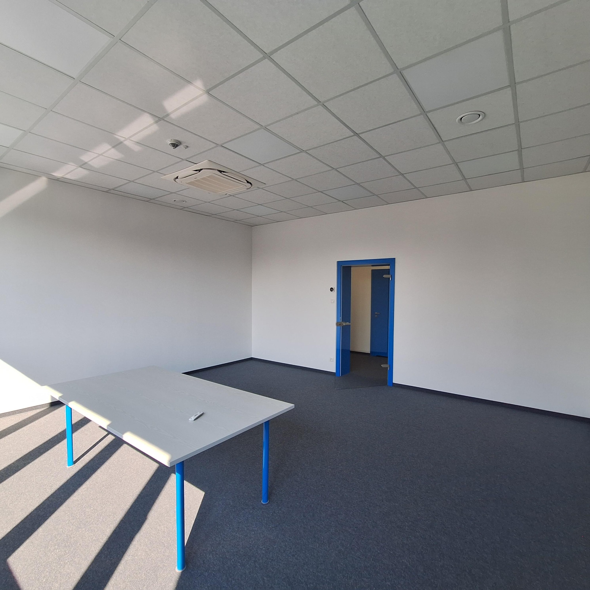 Jasne biuro po remoncie: białe ściany, sufit kasetonowy, szara wykładzina i niebieskie obramowanie drzwi. Stół z niebieskimi nogami w lewym dolnym rogu.