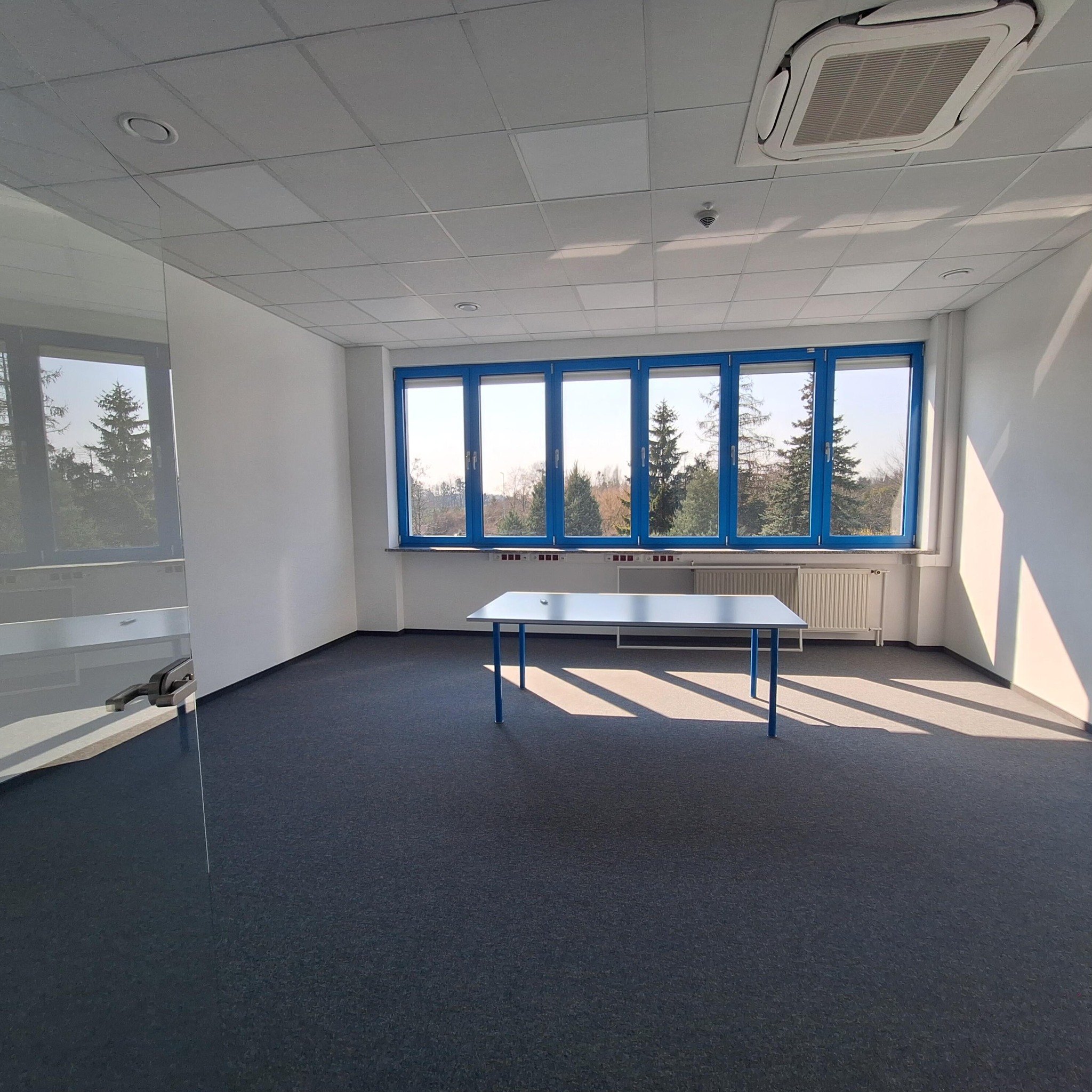 Jasne biuro po remoncie: szara wykładzina, białe ściany, błękitne ramy okien z widokiem na drzewa. Stół z niebieskimi nogami. Klimatyzacja na suficie.