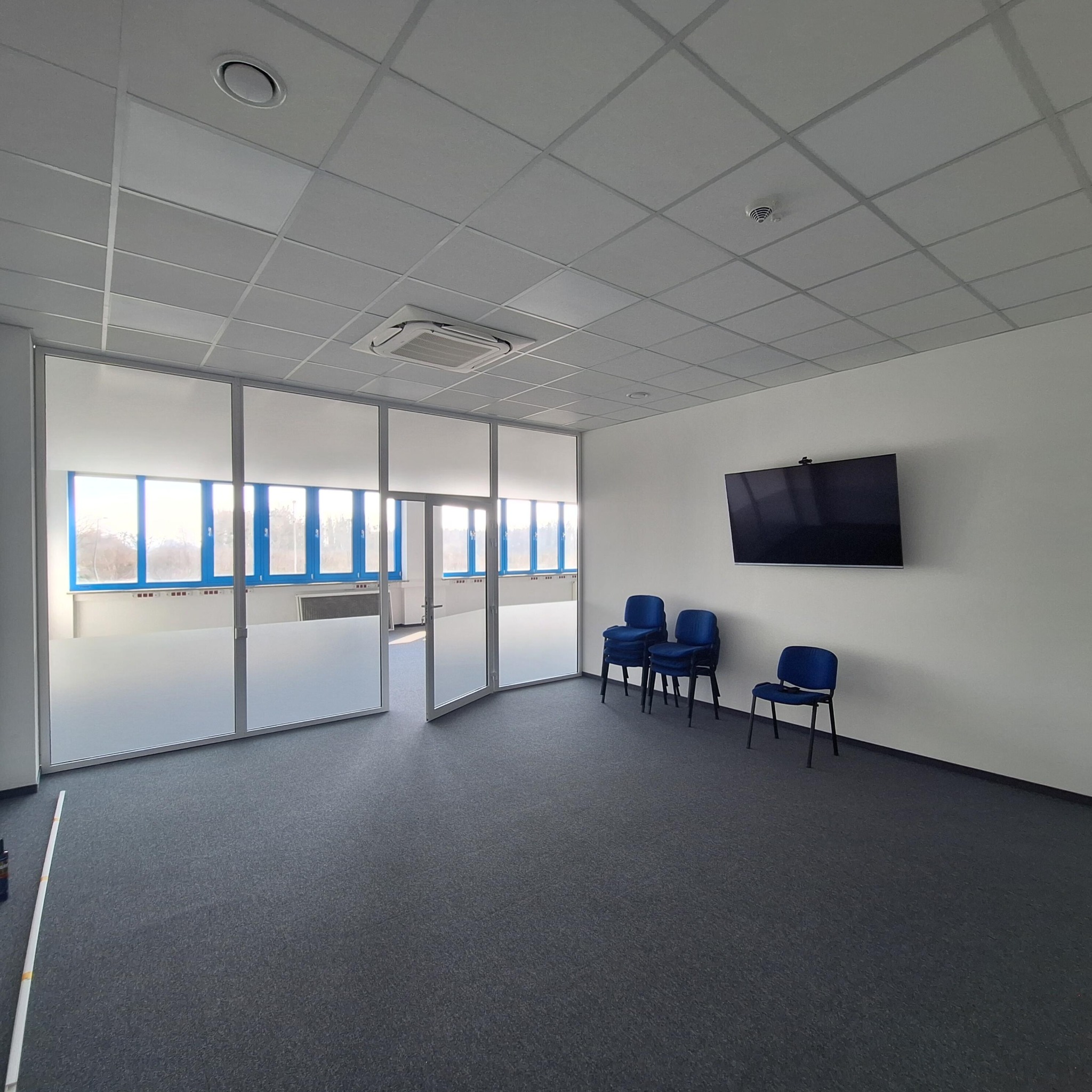 Nowoczesne biuro po remoncie: szklane ściany działowe z matową folią, trzy krzesła i telewizor na białej ścianie. Widoczny sufit podwieszany i szara wykładzina.