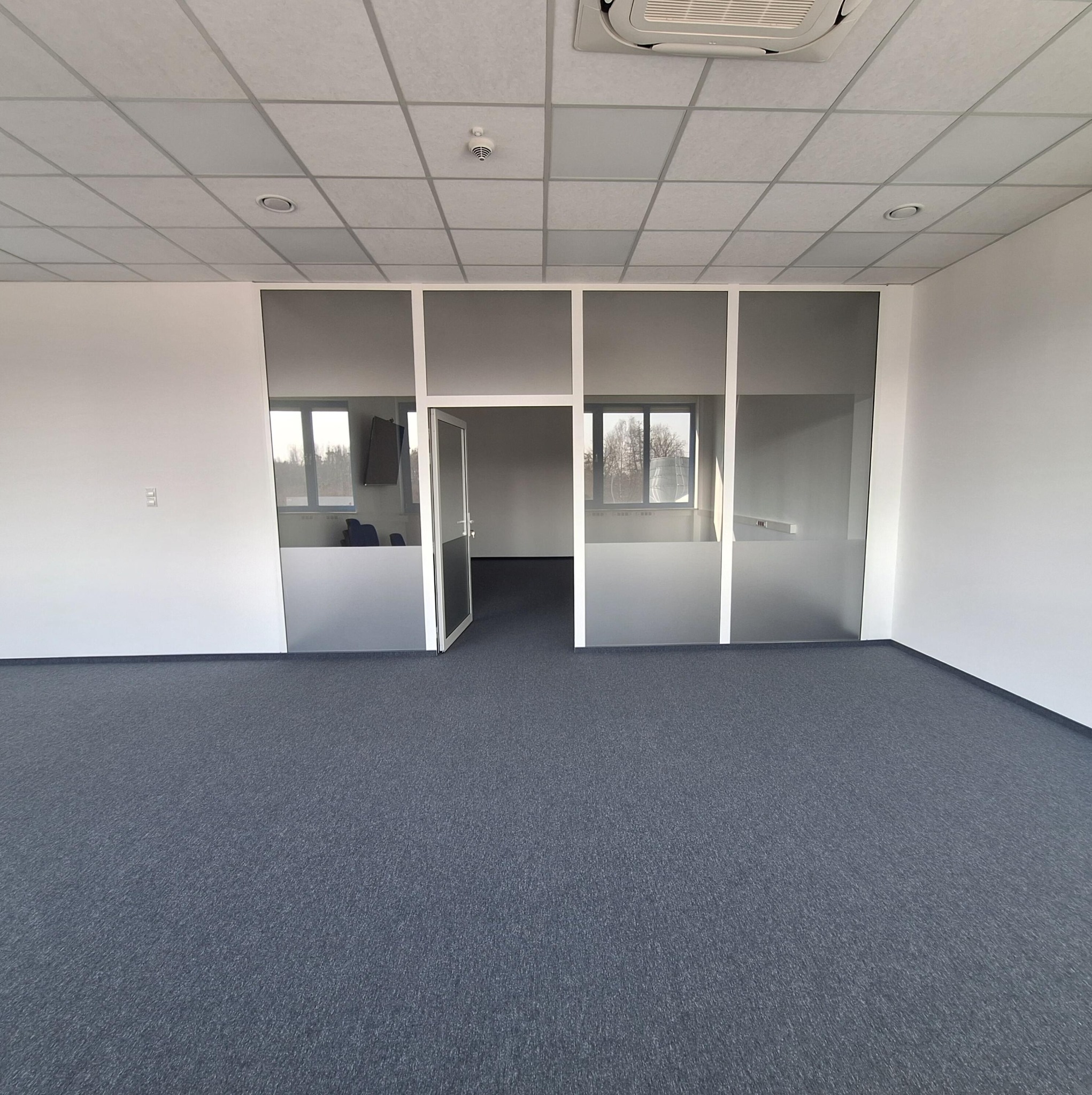 Wykończone wnętrze biurowe z szarą wykładziną i przeszklonymi ściankami działowymi. Widoczny sufit podwieszany i klimatyzacja. Minimalistyczny design.