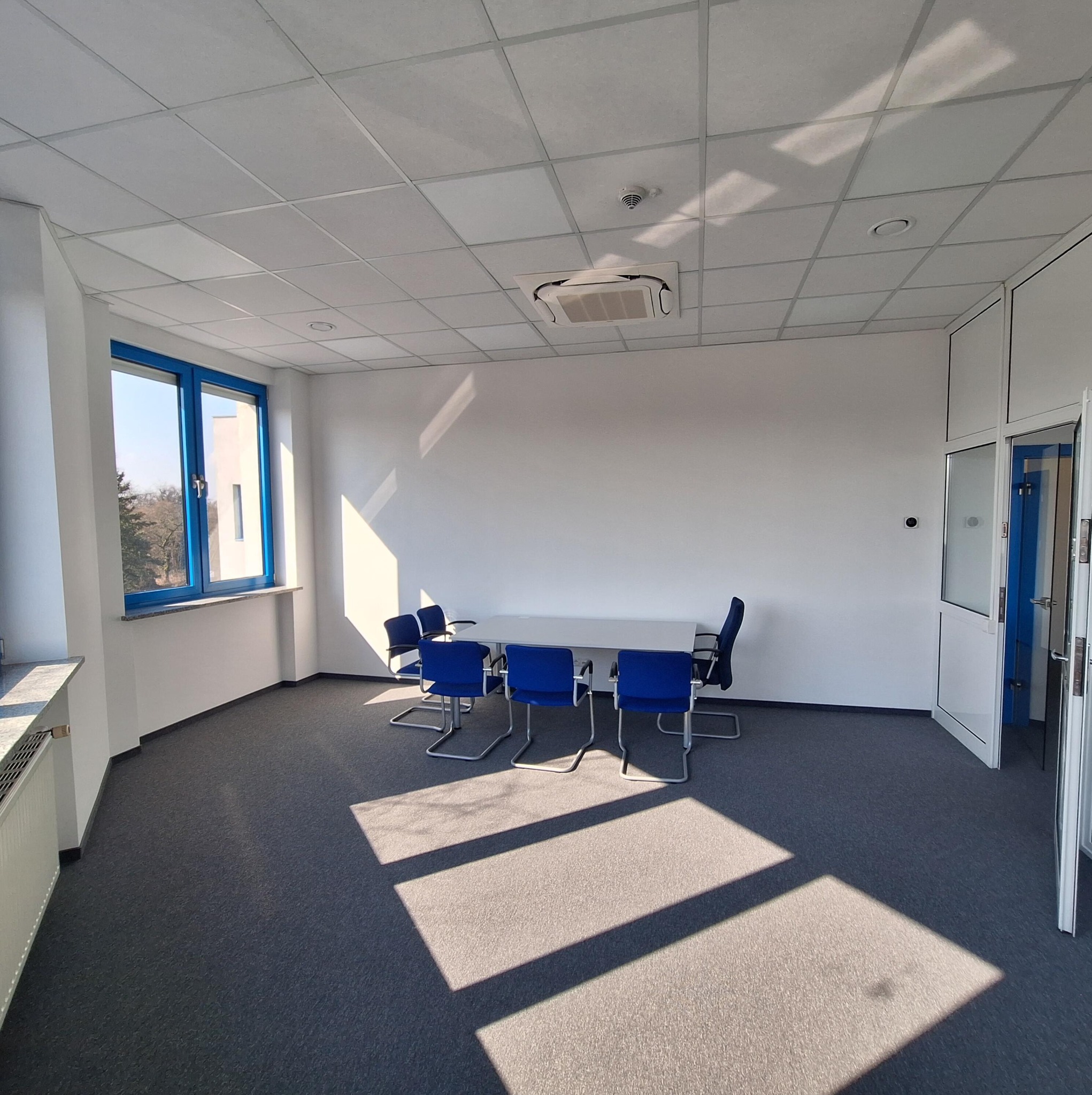 Jasne biuro po remoncie: białe ściany, sufit kasetonowy, okno z niebieską ramą, szara wykładzina i stół konferencyjny z krzesłami. Promienie słoneczne wpadają przez okno.