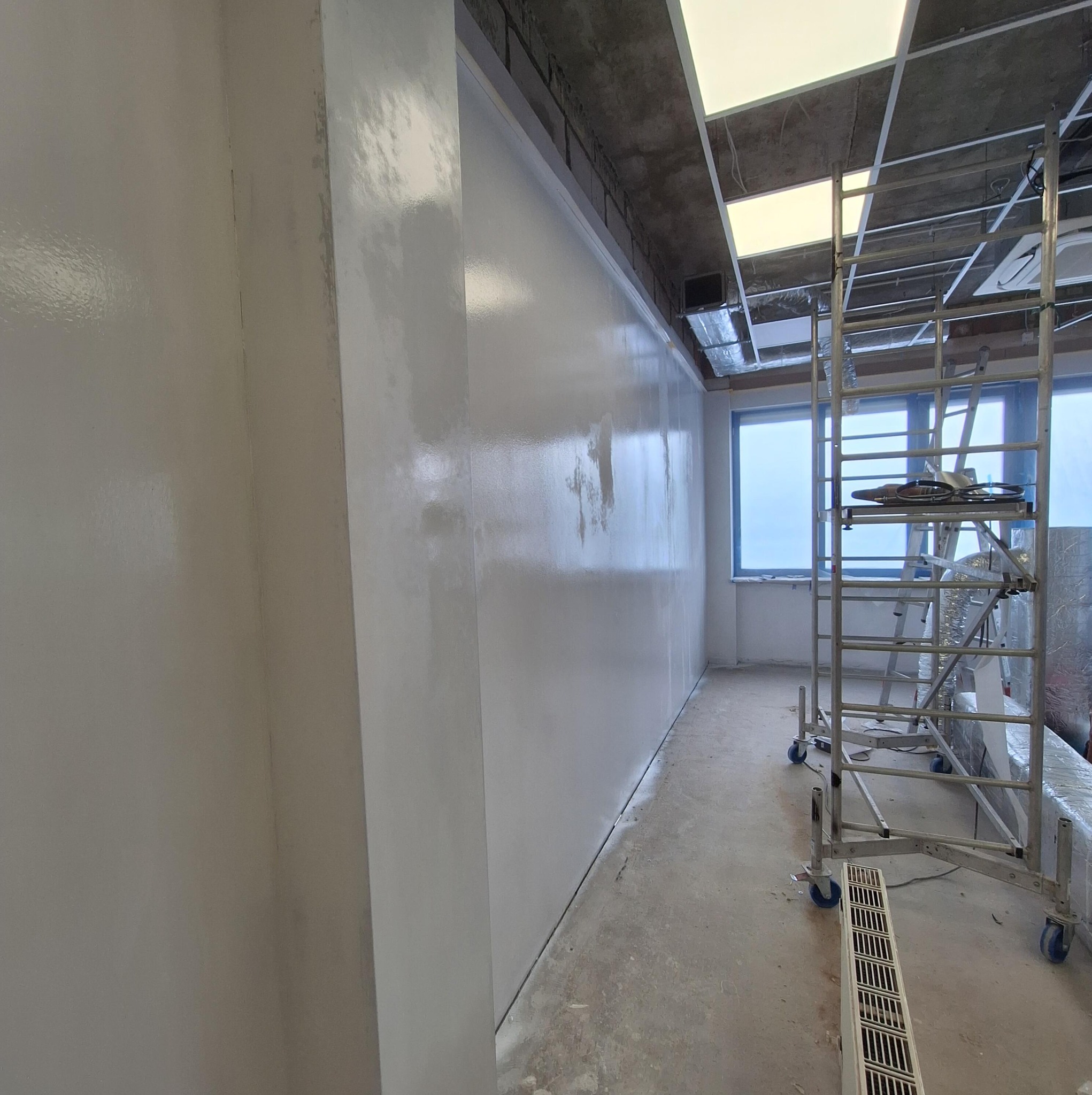 Świeżo pomalowana, błyszcząca ściana w jasnym odcieniu szarości w biurze, z widocznym rusztowaniem i oknem w tle. Widoczne niedoskonałości malowania.
