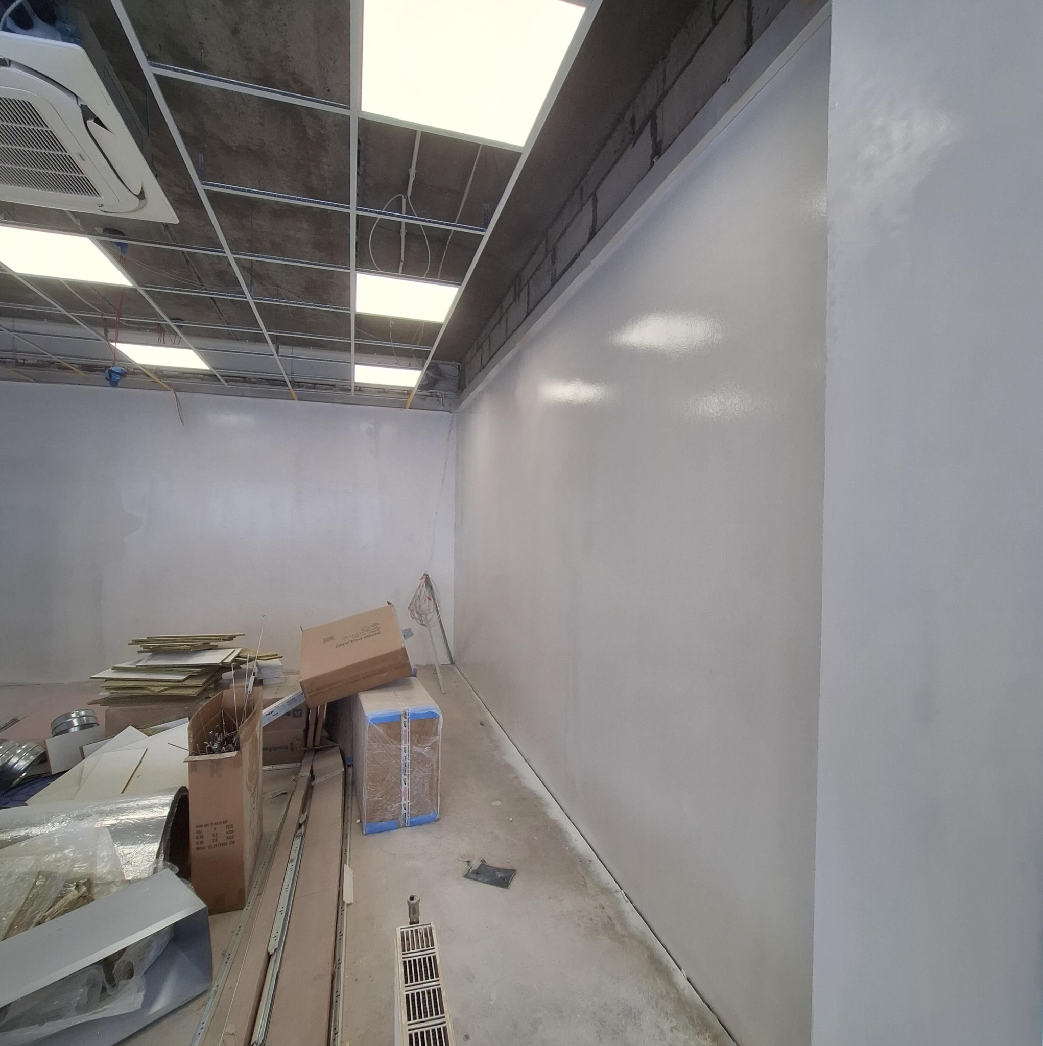 Świeżo pomalowane, gładkie ściany w jasnym odcieniu szarości w pomieszczeniu z surowym stropem i widoczną instalacją. Na podłodze materiały budowlane i kartony.
