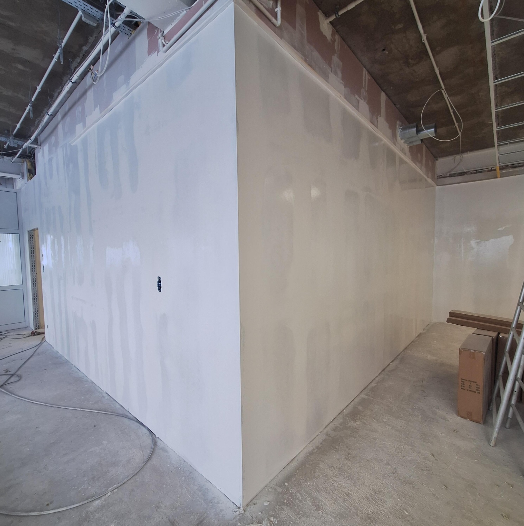 Wnętrze w trakcie prac: ściany po nałożeniu gładzi, widoczne instalacje pod sufitem, surowy beton na podłodze, przygotowane do dalszych prac wykończeniowych.