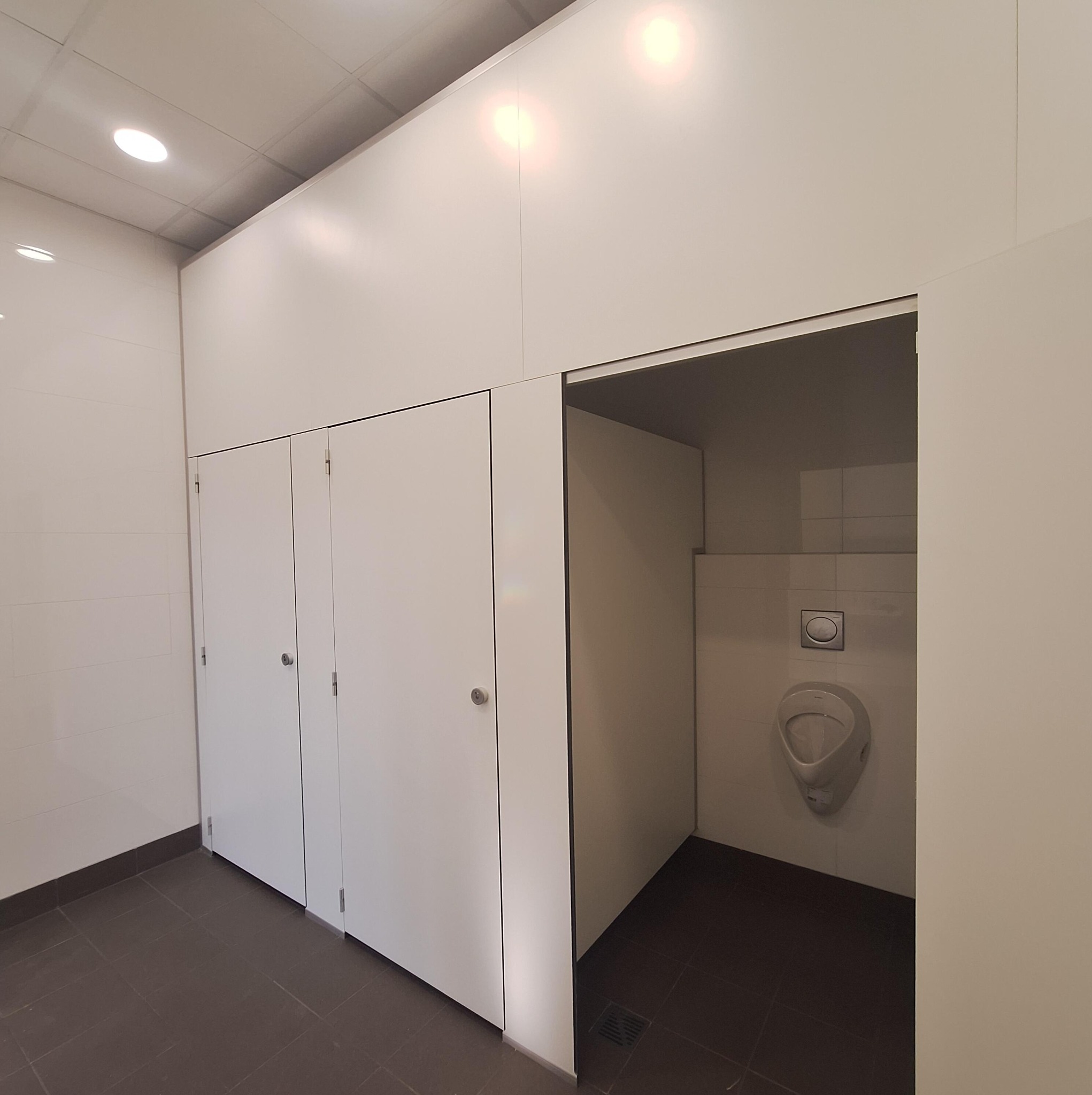 Nowoczesna toaleta publiczna z kabinami i pisuarami, wykończona w bieli, zlokalizowana w Chybach. Minimalistyczny design, ciemna podłoga.