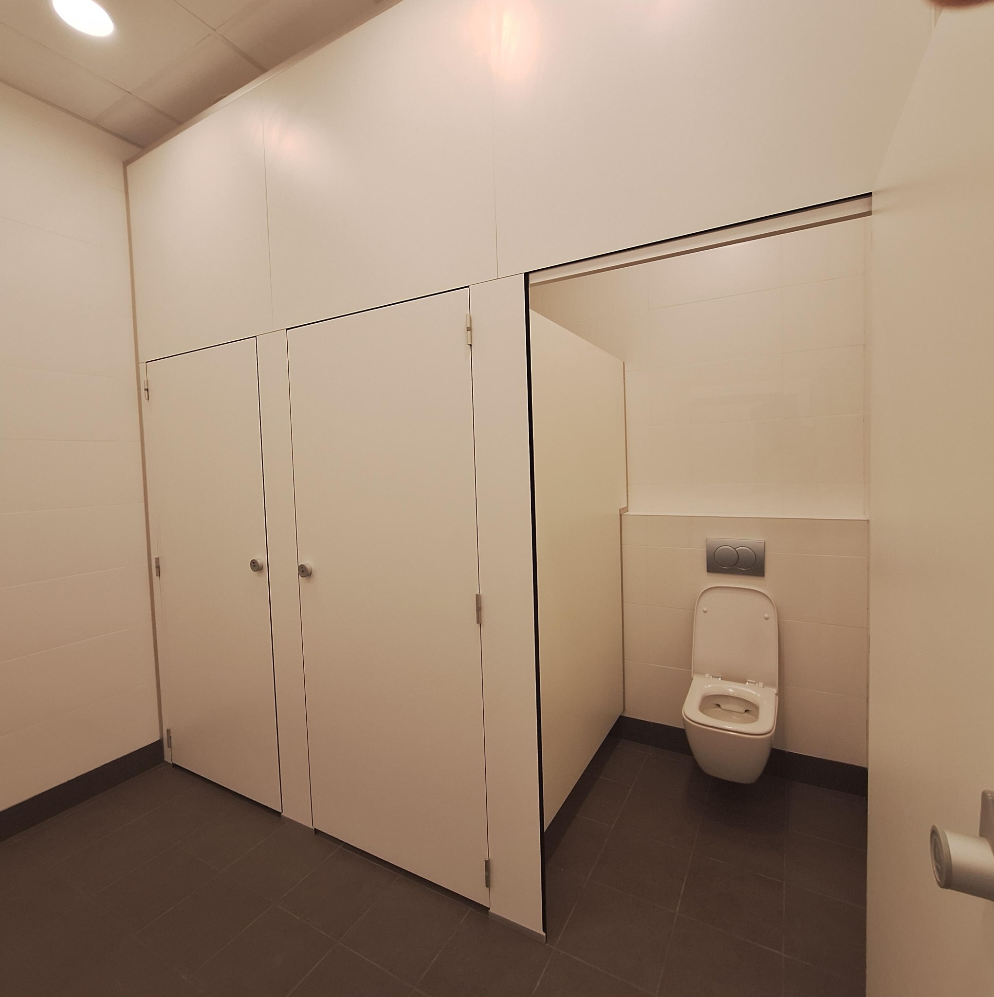 Nowoczesna toaleta publiczna z kabinami i wiszącą muszlą klozetową. Minimalistyczny design, jasne kolory, ciemna podłoga. Widok z perspektywy użytkownika.