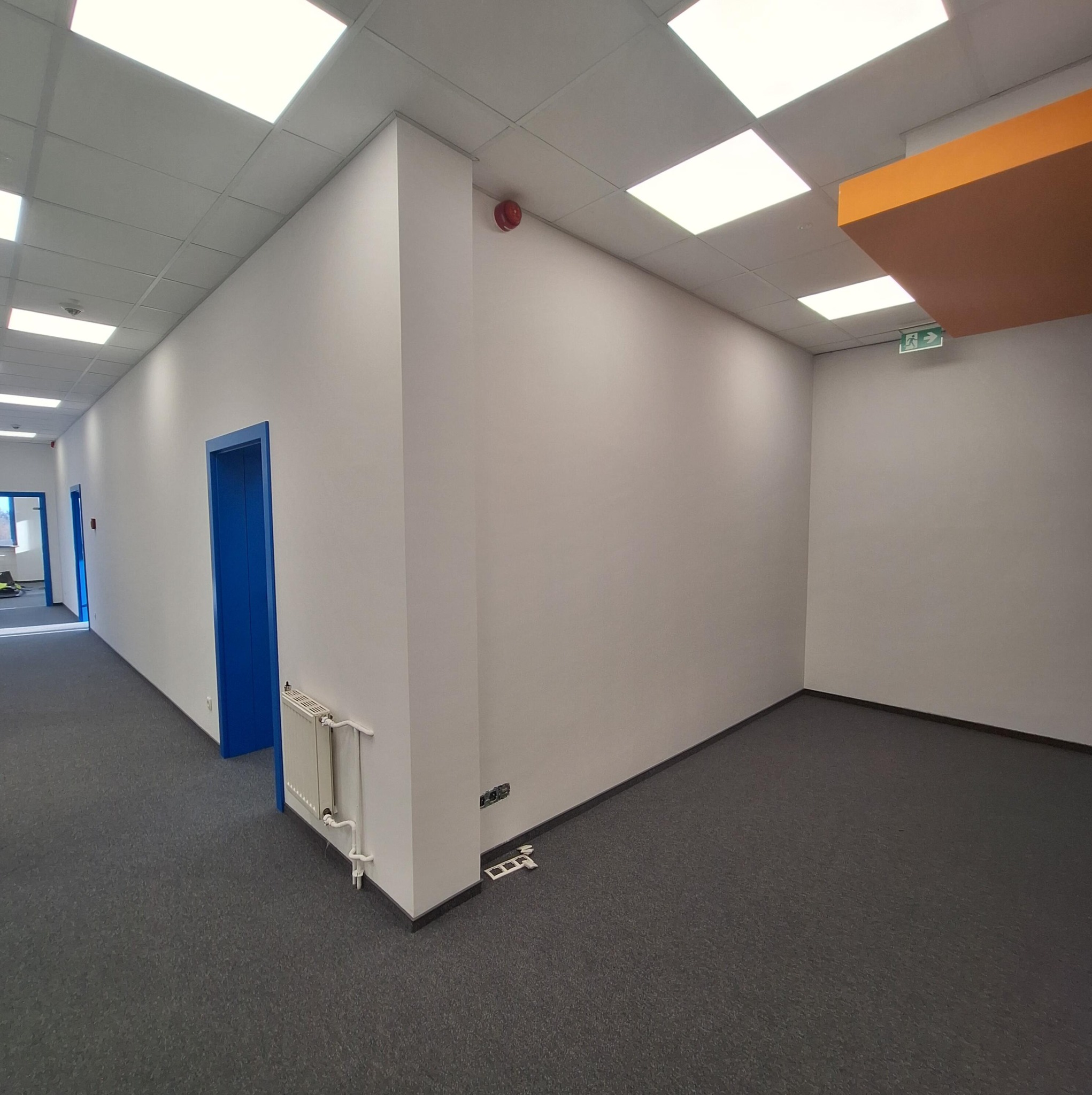 Białe ściany biura z szarą wykładziną, niebieskie ramy drzwi, grzejnik i fragment pomarańczowego sufitu podwieszanego. Widoczne gniazdka elektryczne i czujnik dymu.