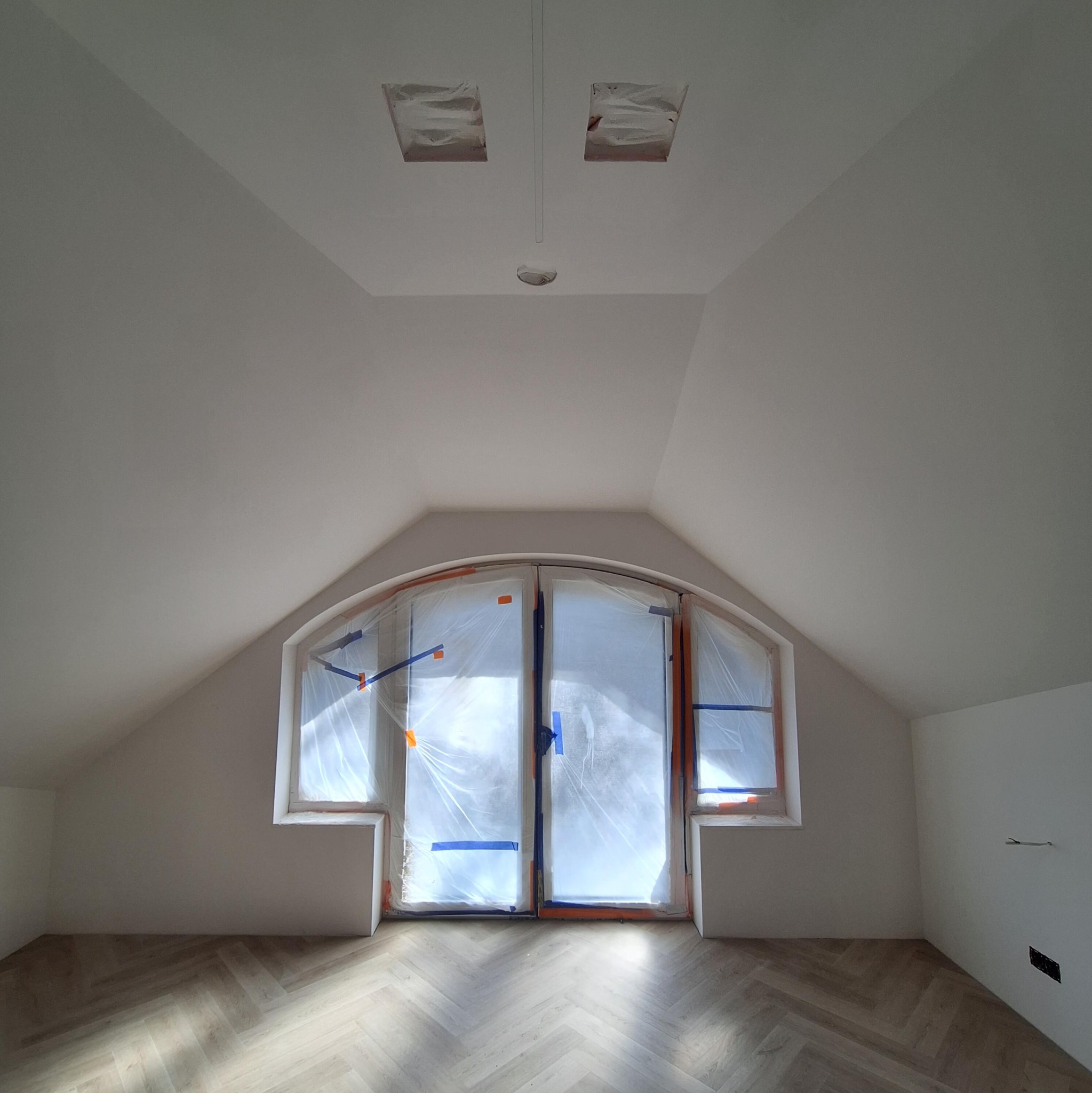 Poddasze po remoncie: świeżo pomalowane ściany i sufit na biało, zabezpieczone okna, parkiet w jodełkę. Widoczne otwory na oświetlenie w suficie.