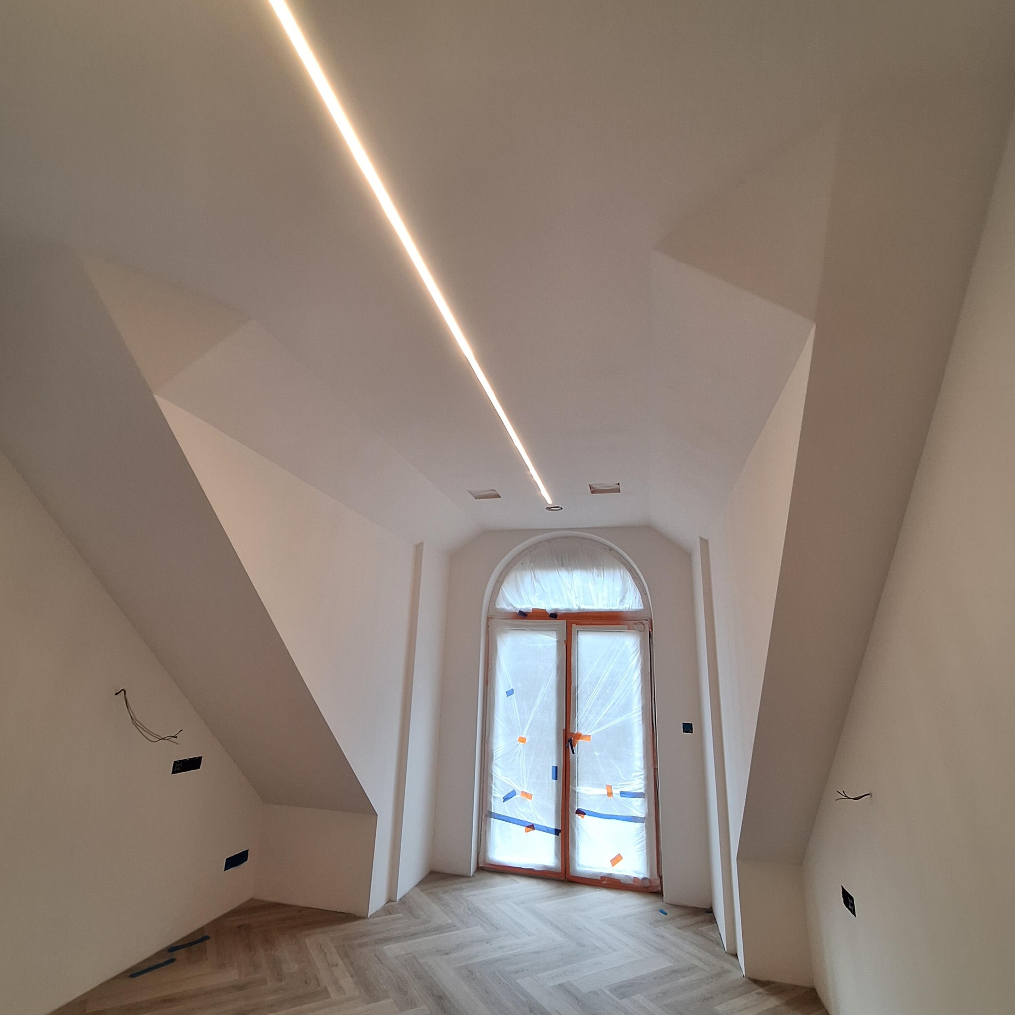 Jasne poddasze z oknem zabezpieczonym folią i drewnianą ramą. Na suficie zamontowane oświetlenie liniowe LED. Parkiet ułożony w jodełkę. Ściany przygotowane do malowania.