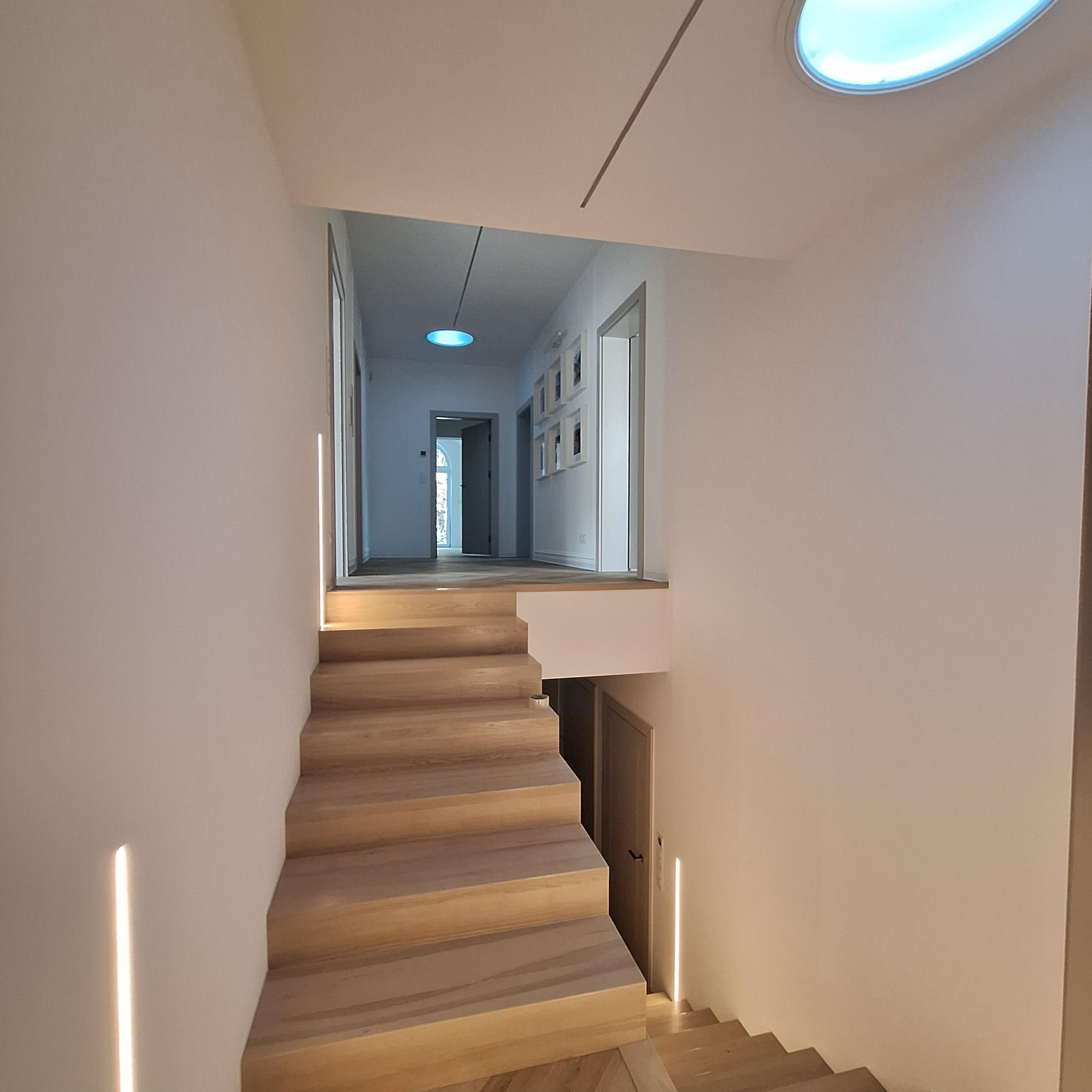 Nowoczesne schody drewniane z subtelnym oświetleniem LED wzdłuż ścian, prowadzące do jasnego korytarza z okrągłym świetlikiem sufitowym i minimalistycznymi dekoracjami.