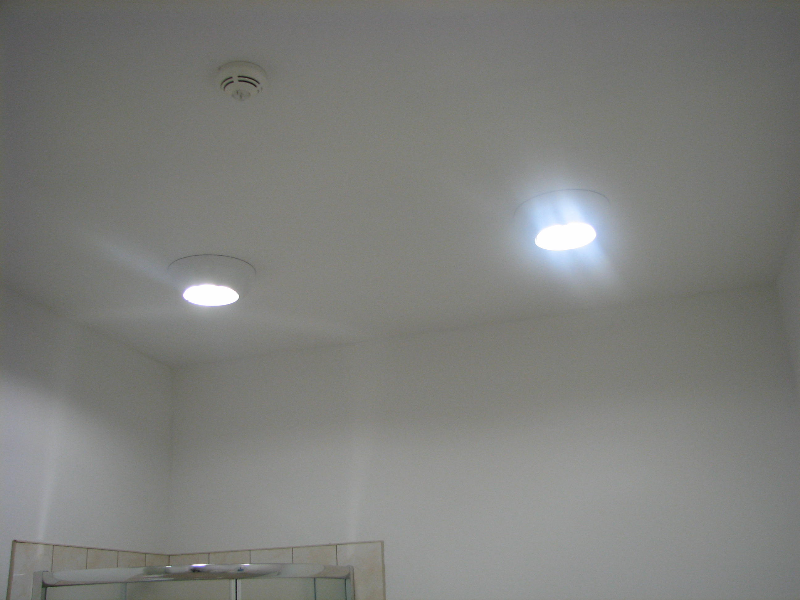 Gładka, świeżo położona, biała ściana z dwoma okrągłymi lampami sufitowymi i czujnikiem dymu. Widoczny fragment kabiny prysznicowej.