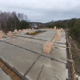 Wylany strop żelbetowy z widocznym zbrojeniem i pustakami ceramicznymi ustawionymi w regularnych odstępach, w tle drzewa i zabudowa jednorodzinna przy pochmurnym niebie.