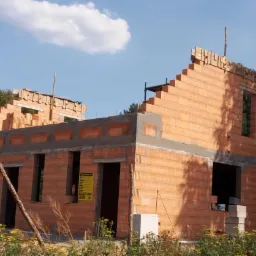 Wznoszenie ścian domu murowanego z bloczków ceramicznych, widoczne nadproża okienne i drzwiowe, częściowo wykończona konstrukcja dachu, tablica informacyjna na pierwszym planie, otoczenie w stanie...