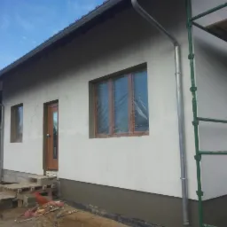 Świeżo otynkowany, jednokondygnacyjny dom w trakcie wykończenia, z zabezpieczonymi oknami folią, widoczne rusztowanie i nieuporządkowany teren wokół budynku.