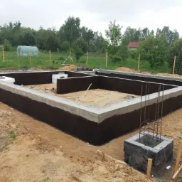 Zalane fundamenty domu jednorodzinnego, zabezpieczone czarną izolacją, z wystającymi zbrojeniami słupów, na piaszczystym terenie działki budowlanej.