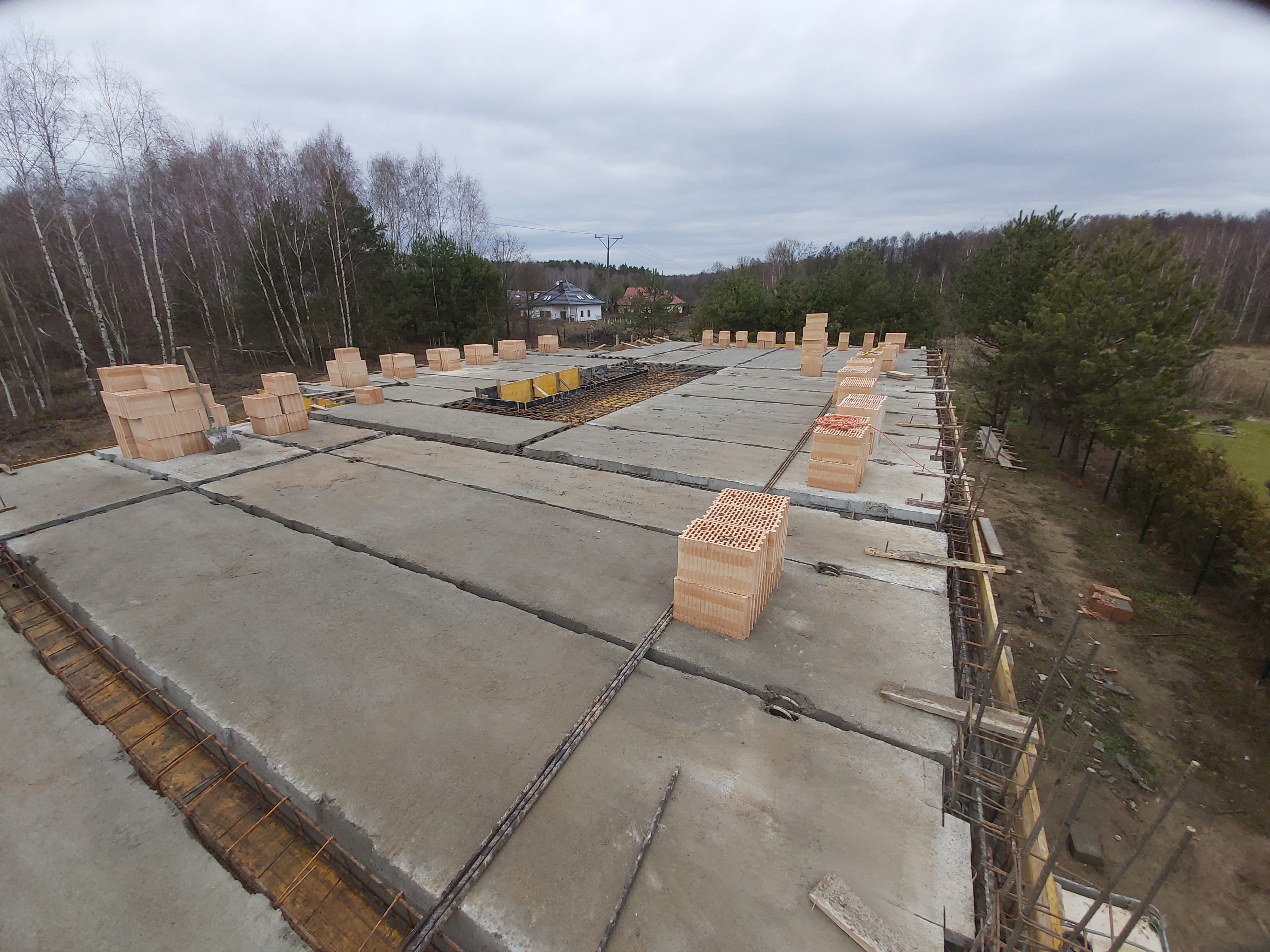 Wylany strop żelbetowy z widocznym zbrojeniem i pustakami ceramicznymi ustawionymi w regularnych odstępach, w tle drzewa i zabudowa jednorodzinna przy pochmurnym niebie.