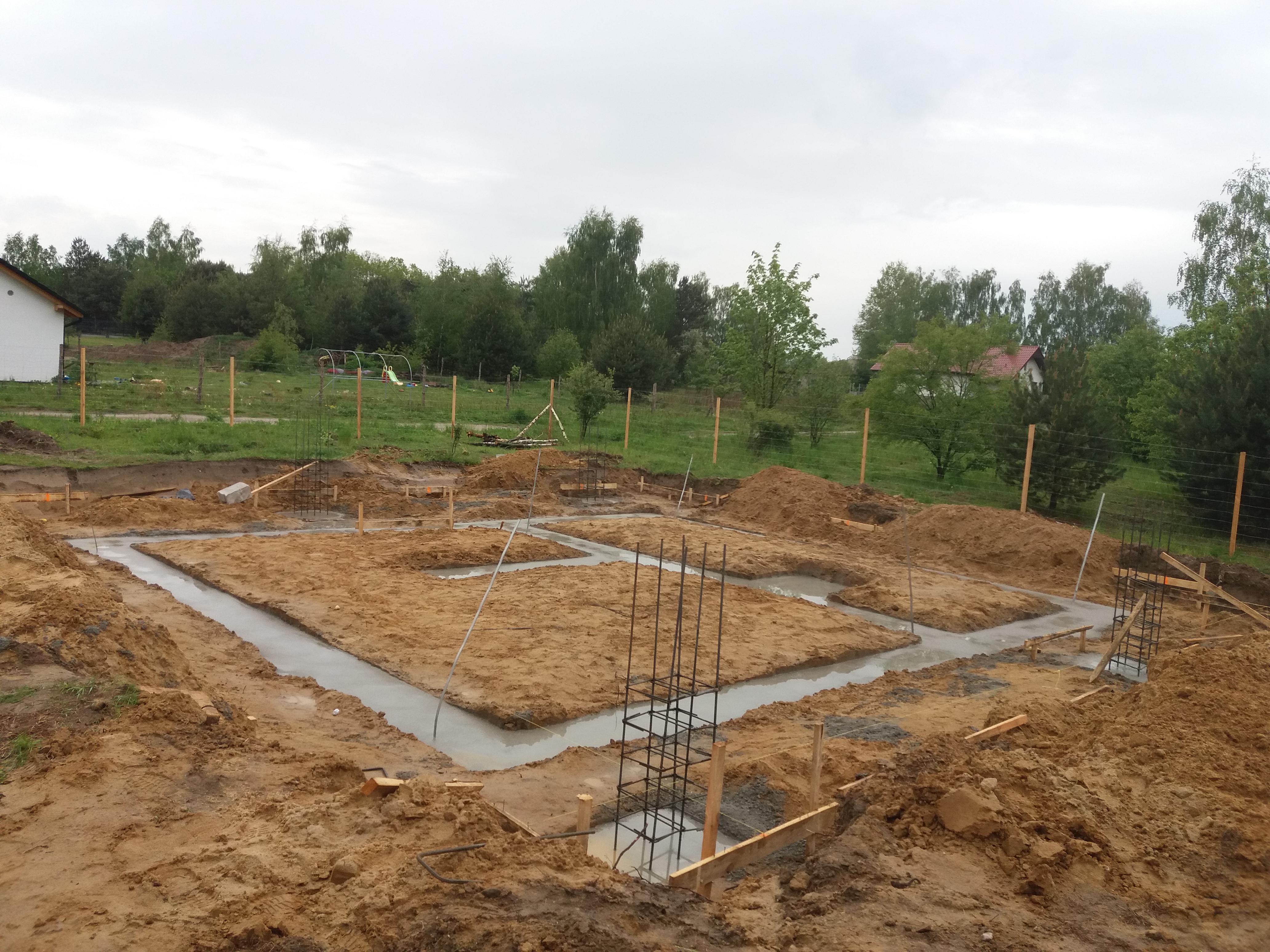 Wylane fundamenty pod dom jednorodzinny, widoczne zbrojenia i szalunki, teren przygotowany pod budowę, w tle drzewa i częściowo widoczny budynek.