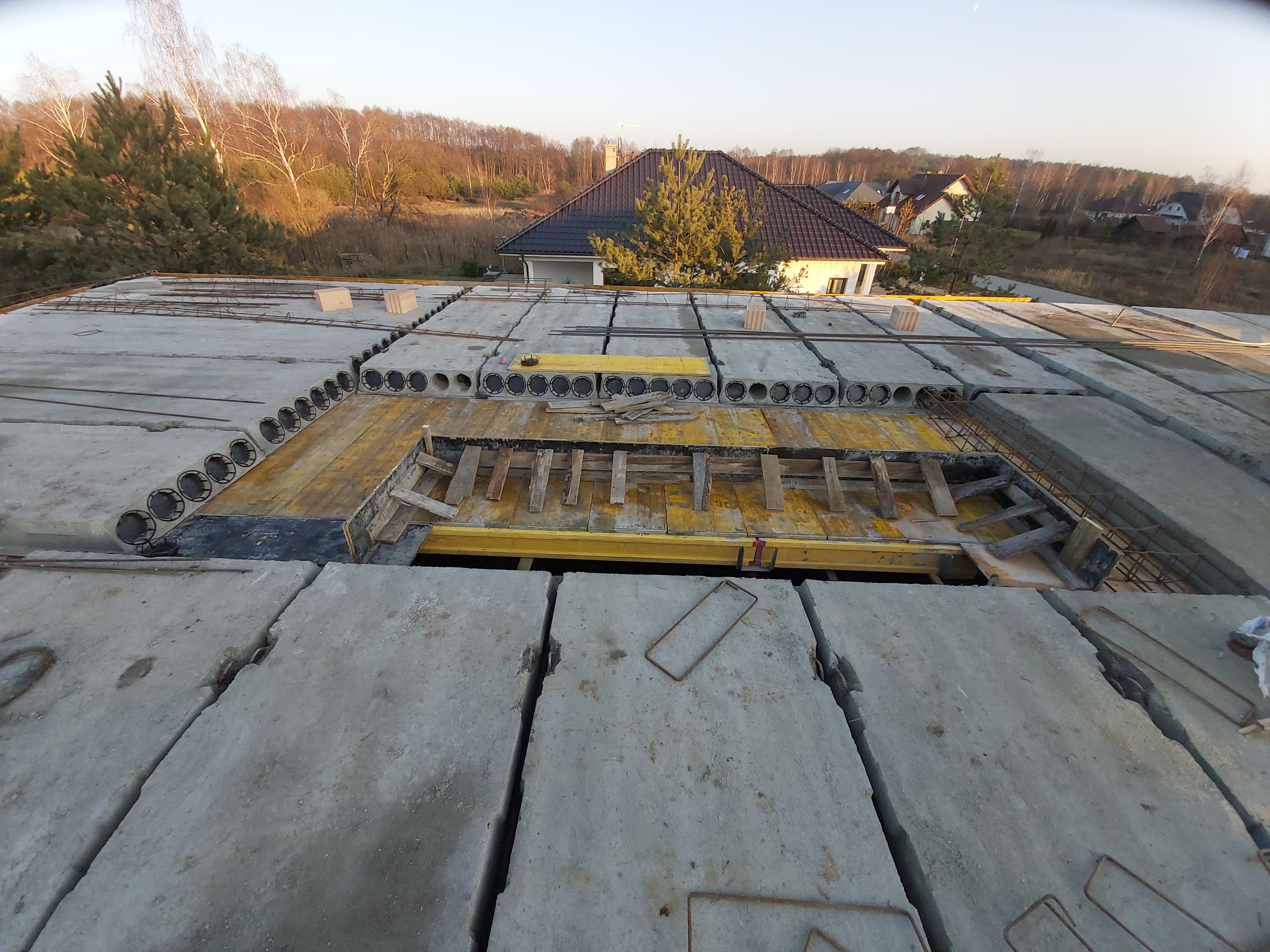 Widok z góry na strop monolityczny z prefabrykatów, przygotowany do zalania betonem; widoczna szalunek z żółtymi elementami i zbrojenie, w tle budynek mieszkalny w zabudowie jednorodzinnej i drzewa.