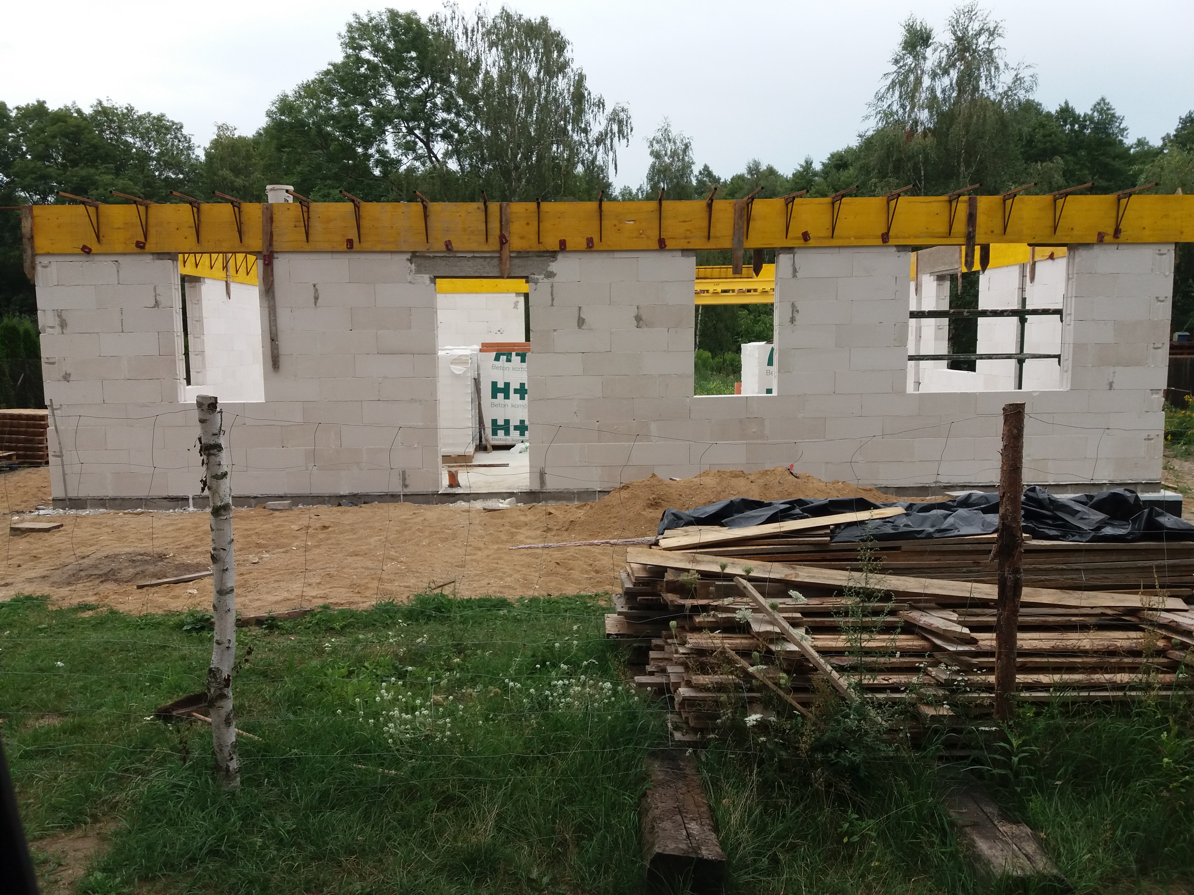 Surowy stan budynku z białych bloczków, widoczne żółte stemple podtrzymujące nadproża, w tle palety z bloczkami H+H, ujęcie zza prowizorycznego ogrodzenia z gałęzi brzozy.