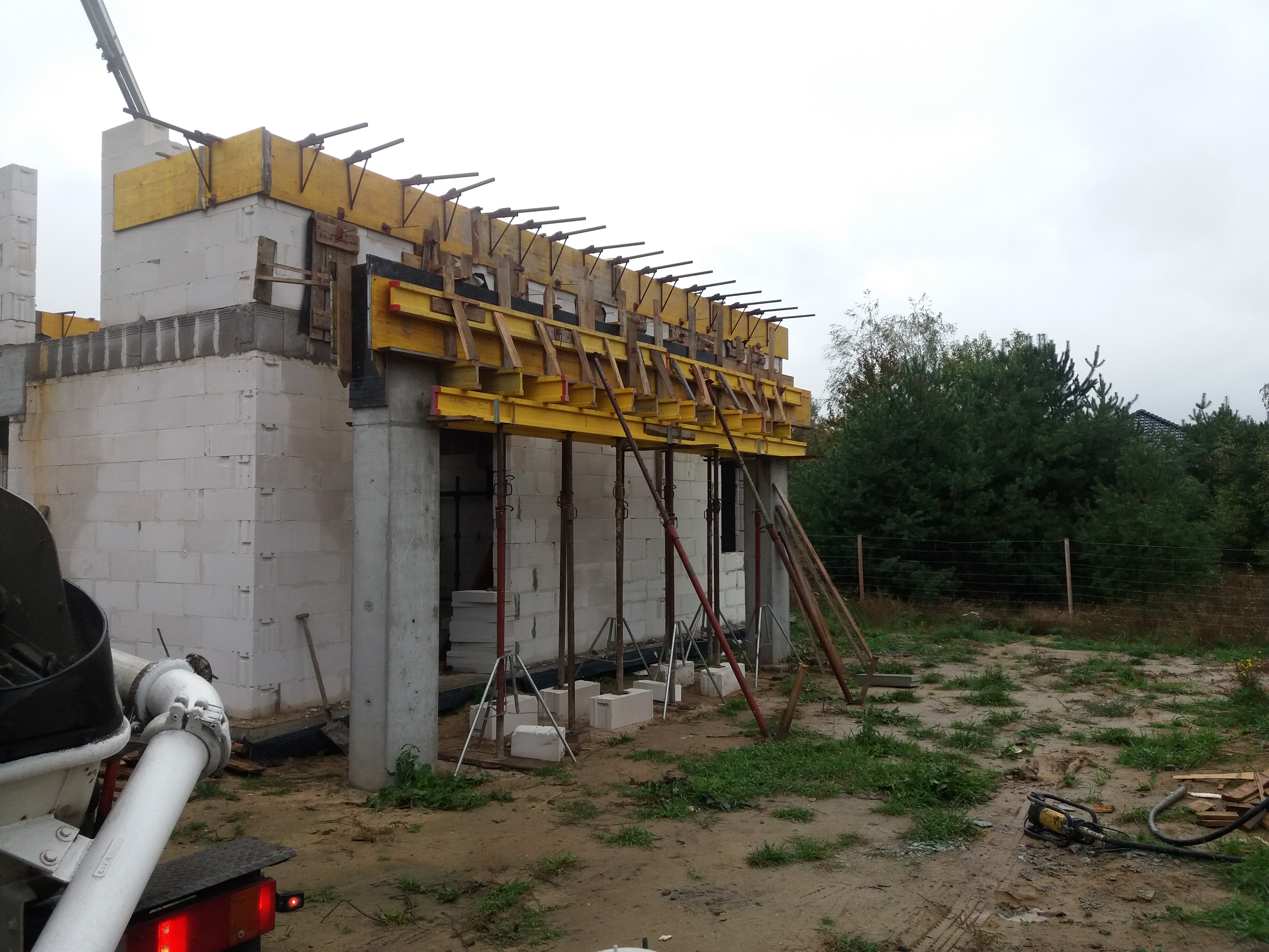 Budowa domu z białych bloczków, widoczne żółte szalunki na stropie i betonowy mikser w tle, teren budowy z trawą i drzewami.