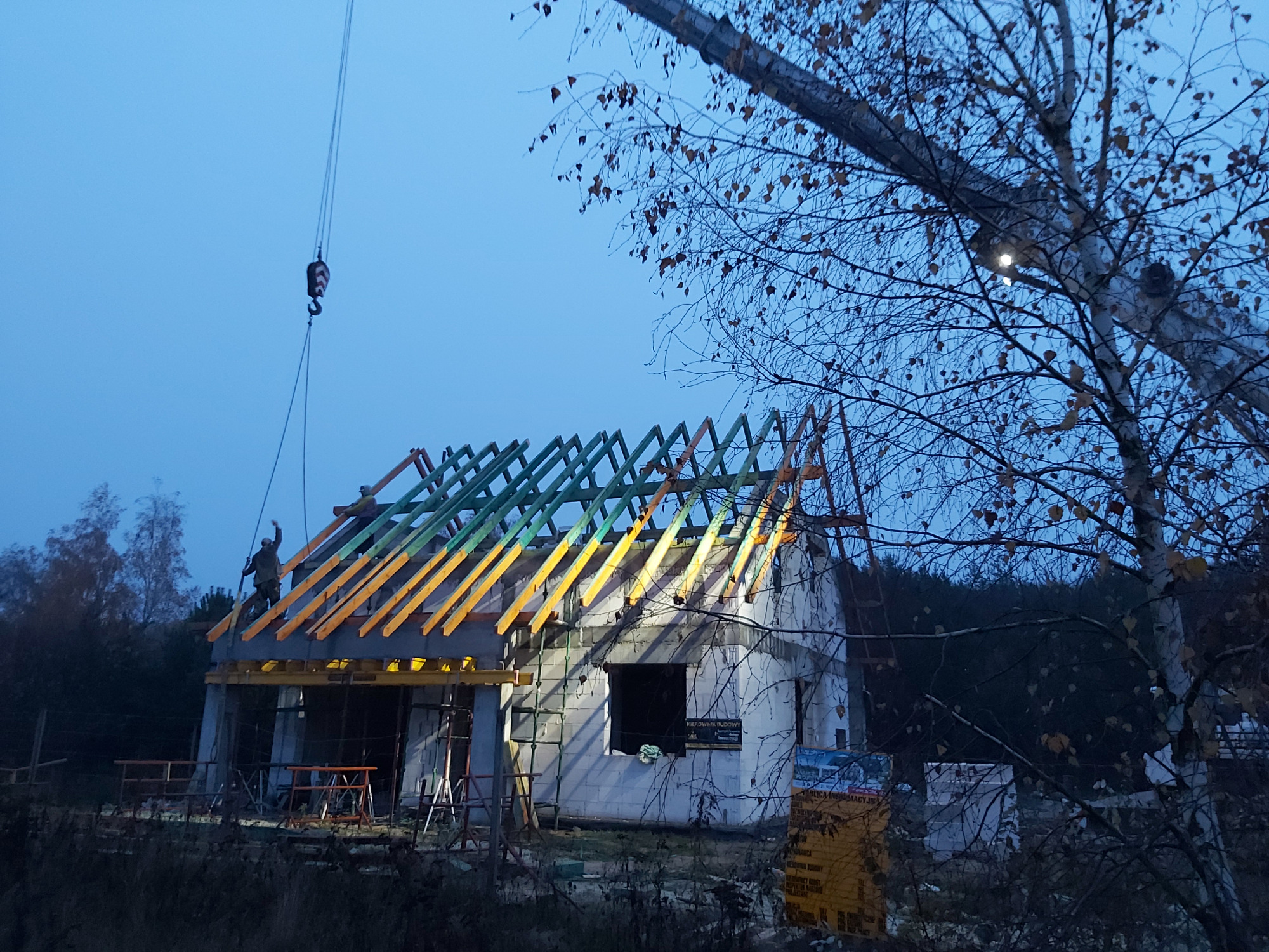 Wczesnym rankiem, budowa domu z pustaków: widoczny szkielet dachu z drewnianych belek w kolorze żółtym i zielonym, pracownicy na dachu, żuraw budowlany w tle, drzewa bez liści na pierwszym planie.