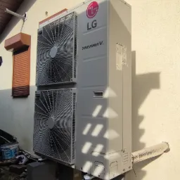 Zewnętrzna jednostka LG Therma V, zamontowana na betonowych blokach przy ścianie budynku z widoczną elewacją i brązową roletą okienną.