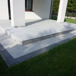 Granitowy taras wejściowy z dwoma stopniami, wykończony ciemniejszą kostką brukową, przed białym domem z kolumnami i zadbanym trawnikiem.