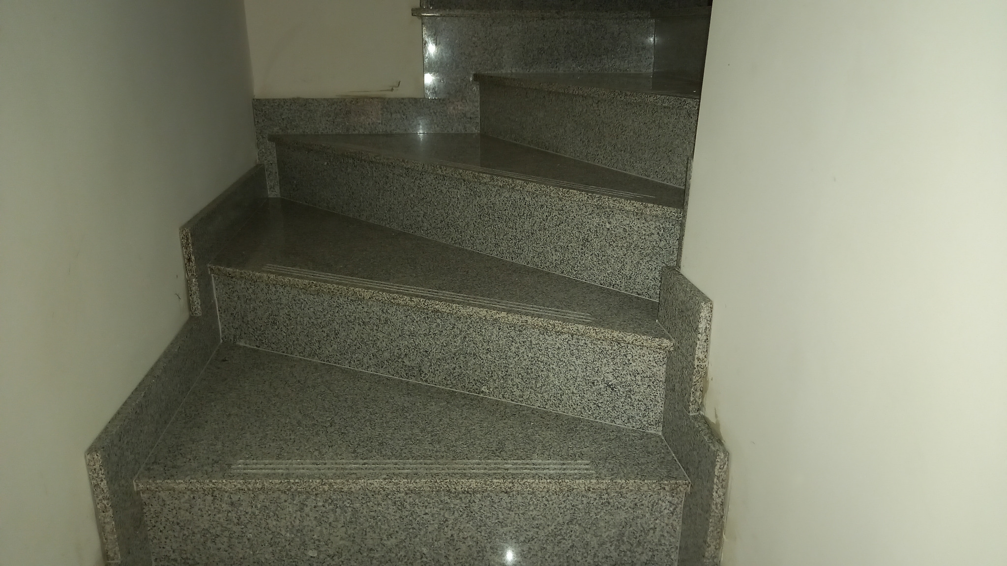 Klatka schodowa z szarymi, kamiennymi stopniami, widok z góry na zakręcające schody wewnątrz budynku oświetlonych słabym światłem.