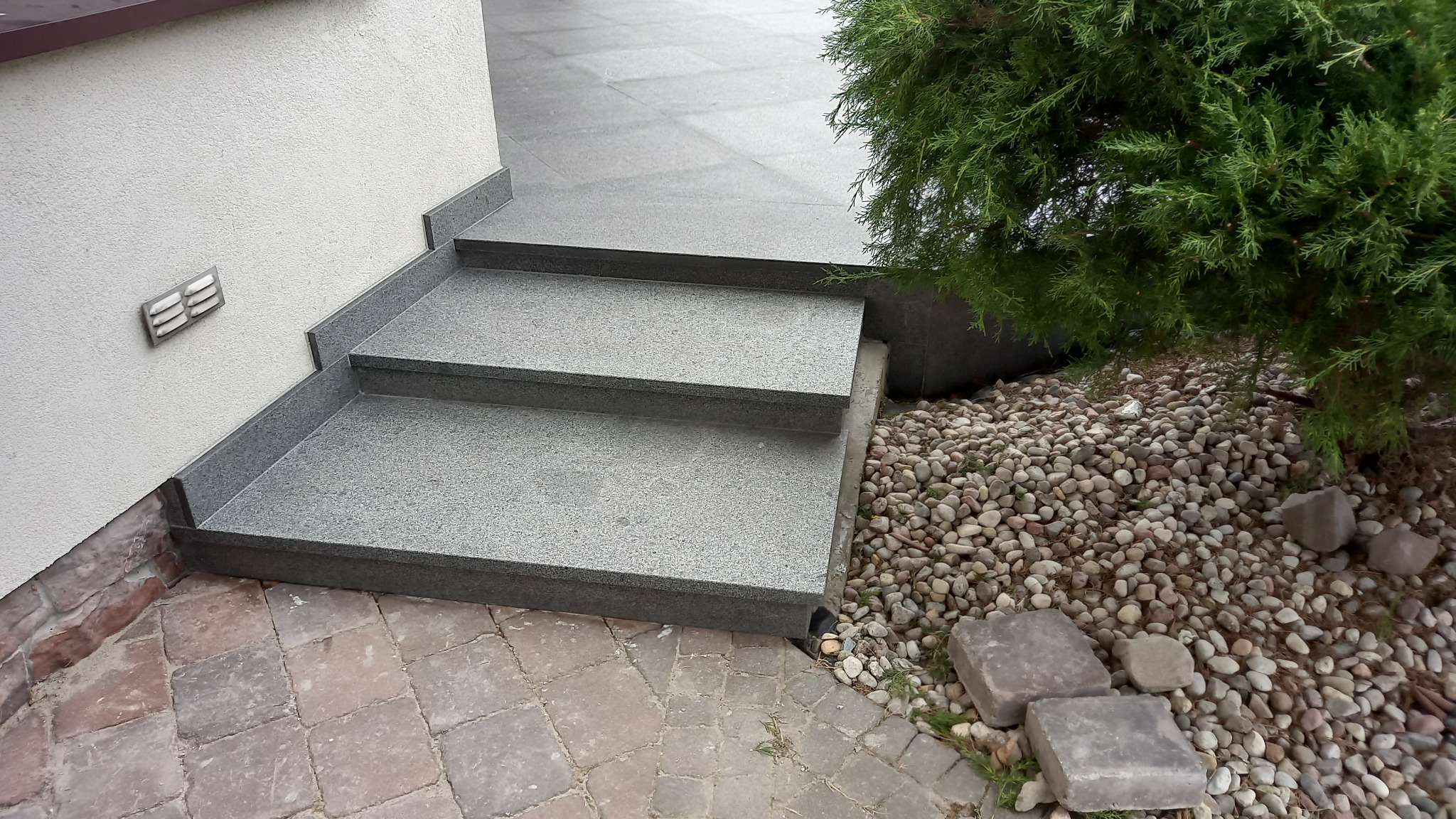 Dwu-stopniowe schody zewnętrzne z szarego, nakrapianego kamienia, przylegające do jasnej ściany budynku z kratką wentylacyjną, obok iglastego krzewu i wysypanych kamyków.