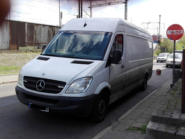 Biały samochód dostawczy Mercedes-Benz Sprinter zaparkowany na ulicy miejskiej z chodnikiem i znakiem drogowym w tle.