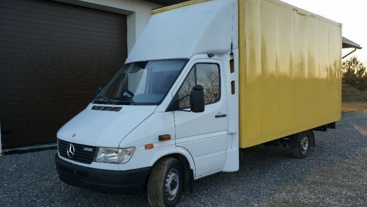 Biały samochód dostawczy Mercedes-Benz Sprinter z żółtą zabudową kontenerową, zaparkowany na żwirowym podjeździe przed budynkiem z brązową bramą garażową.